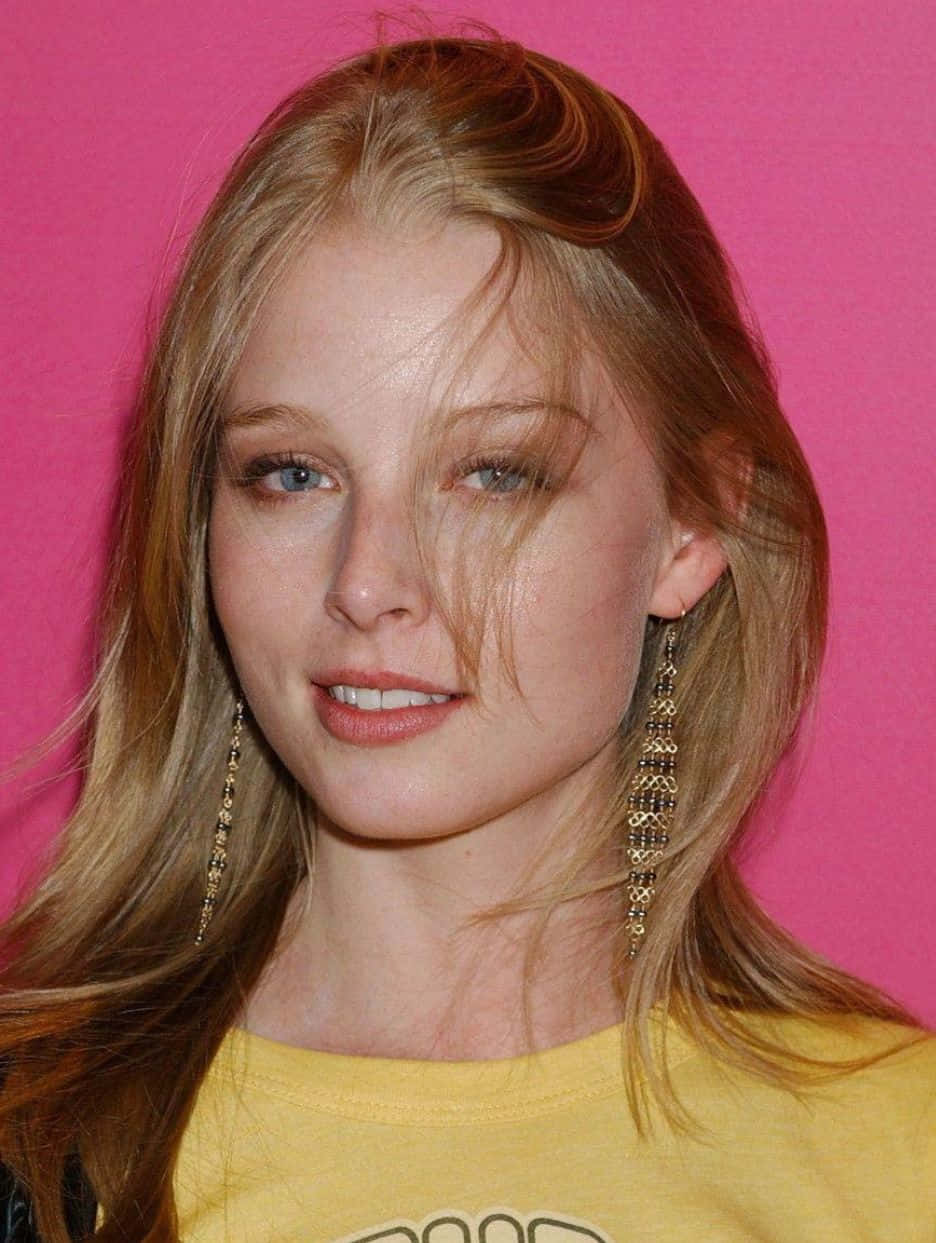 rachel nichols 桌布 壁紙 — 一位金髮藍眼的女士在鮮豔的粉紅色背景前微笑，穿著黃色襯衫和長串珠耳環。