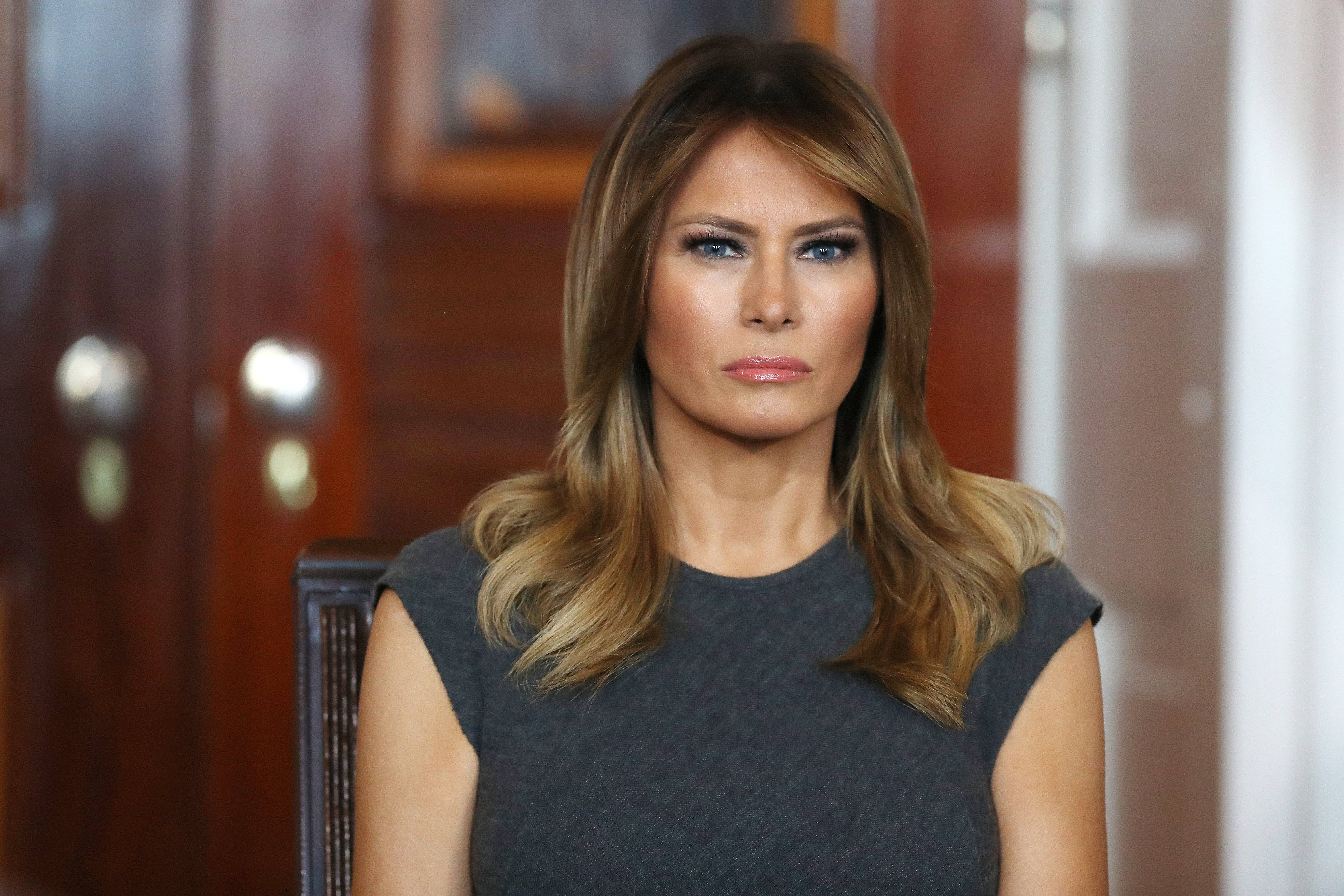Melania 桌布 壁紙 — 一位金髮藍眼的女士坐在木椅上，身穿深灰色上衣，背景是模糊的木門。