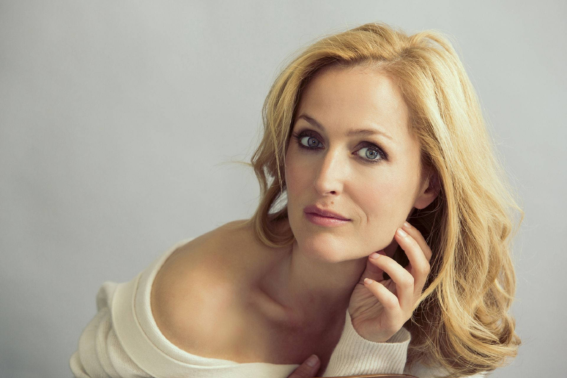 gillian anderson 桌布 壁紙 — 一位金髮藍眼的女性，在純灰色背景前，將手靠近臉頰，若有所思地凝視著。