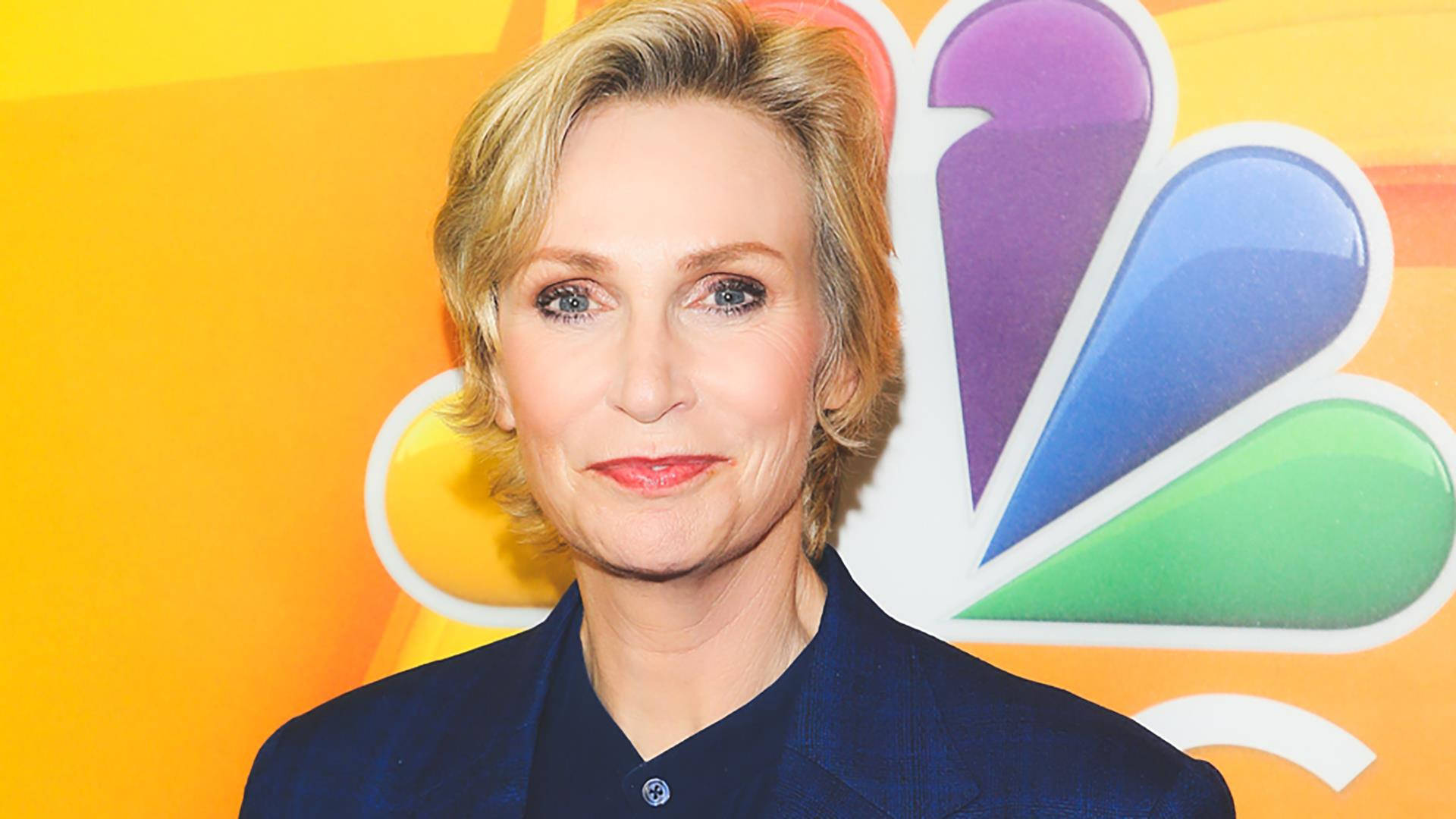 Celebrity 桌布 壁紙 — 一位金髮藍眼的女性，在色彩繽紛的NBC孔雀標誌背景前，穿著深藍色西裝外套。