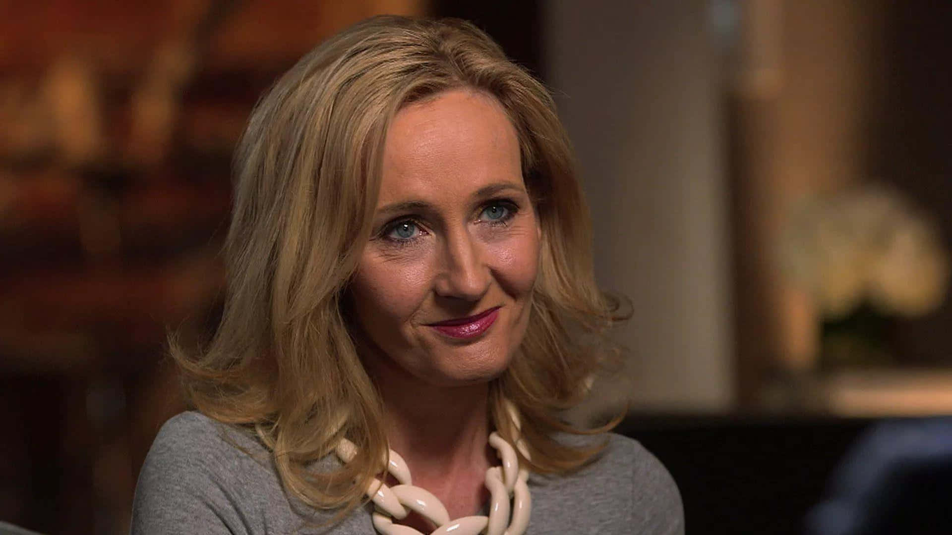 J K Rowling 桌布 壁紙 — 一位金髮藍眼的女性，穿著灰色上衣和奶油色項鍊，溫暖地微笑。