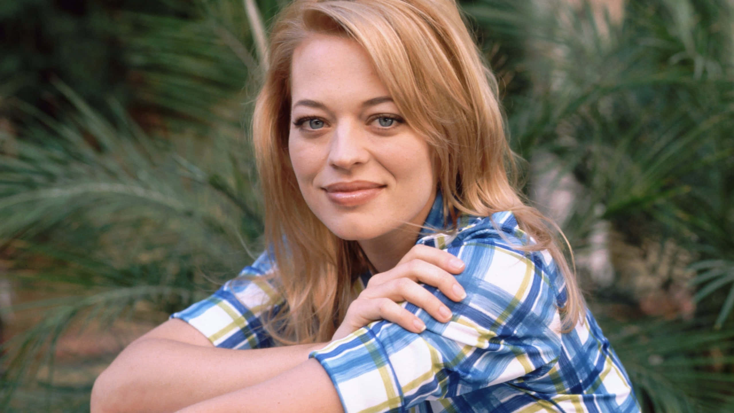 Jeri Ryan 桌布 壁紙 — 一位金髮藍眼的女性，穿著藍色格紋襯衫，在模糊的綠色棕櫚樹背景下溫柔微笑。