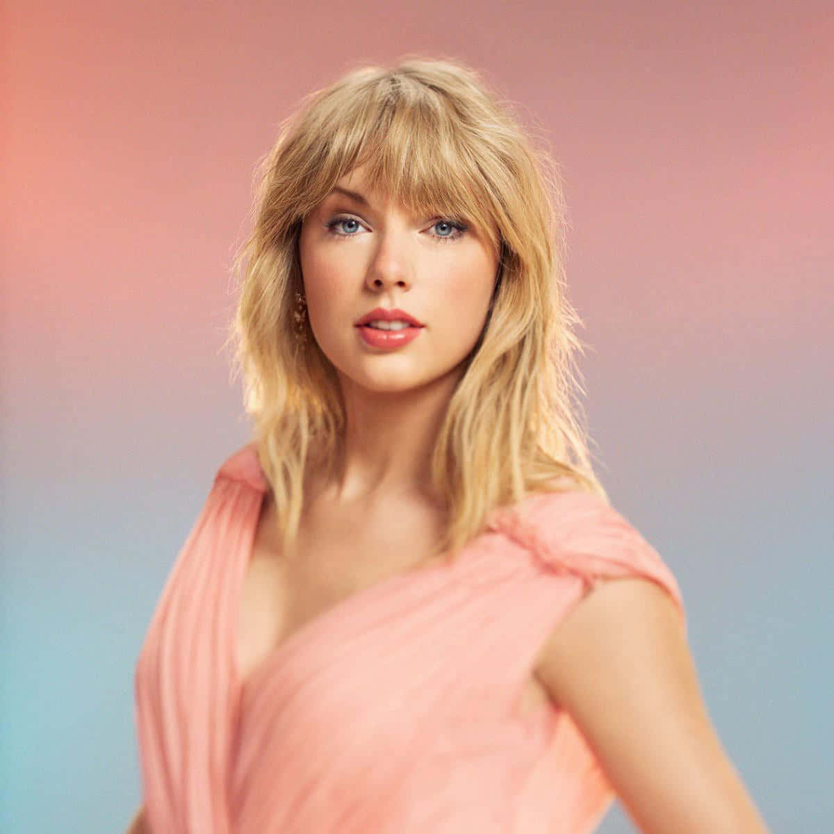Taylor Swift Lover 桌布 壁紙 — 一位金髮藍眼的女性，身穿柔軟的粉紅色垂褶上衣，背景為粉紅和藍色漸變。