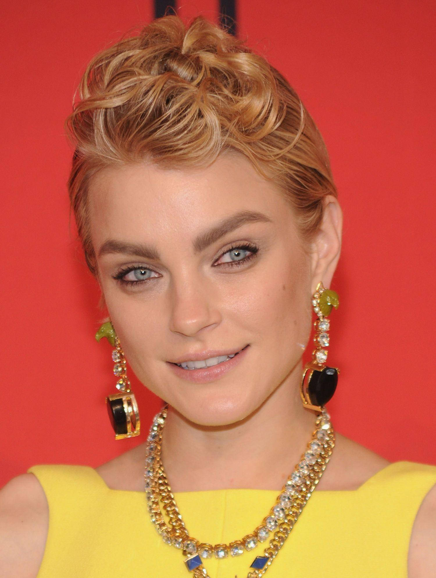 jessica stam 桌布 壁紙 — 一位金髮藍眼的女性在鮮紅色背景前微笑，身穿黃色連衣裙和華麗珠寶。