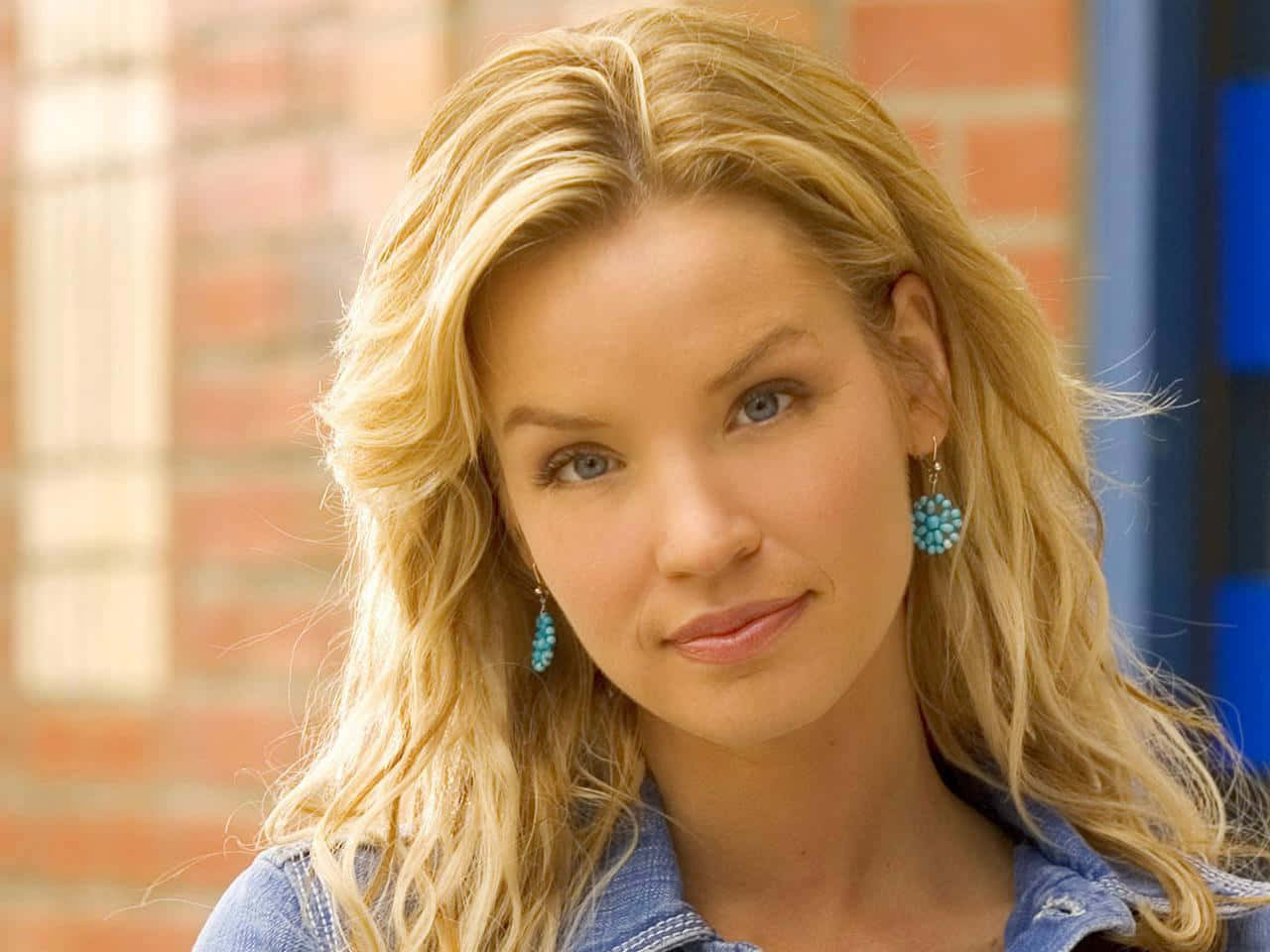 Ashley Scott 桌布 壁紙 — 一位金髮藍眼的女性戴著綠松石耳環和牛仔襯衫，在磚牆背景下溫柔微笑。