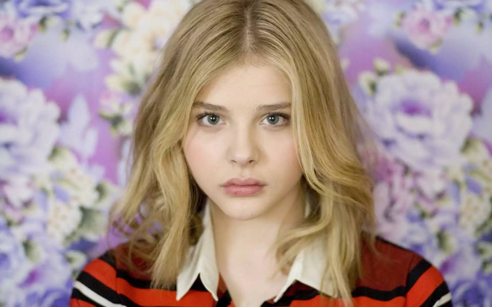 Chloë Grace Moretz 桌布 壁紙 — 一位金髮藍眼的年輕女子在充滿活力的紫色花卉壁紙背景下直視鏡頭。