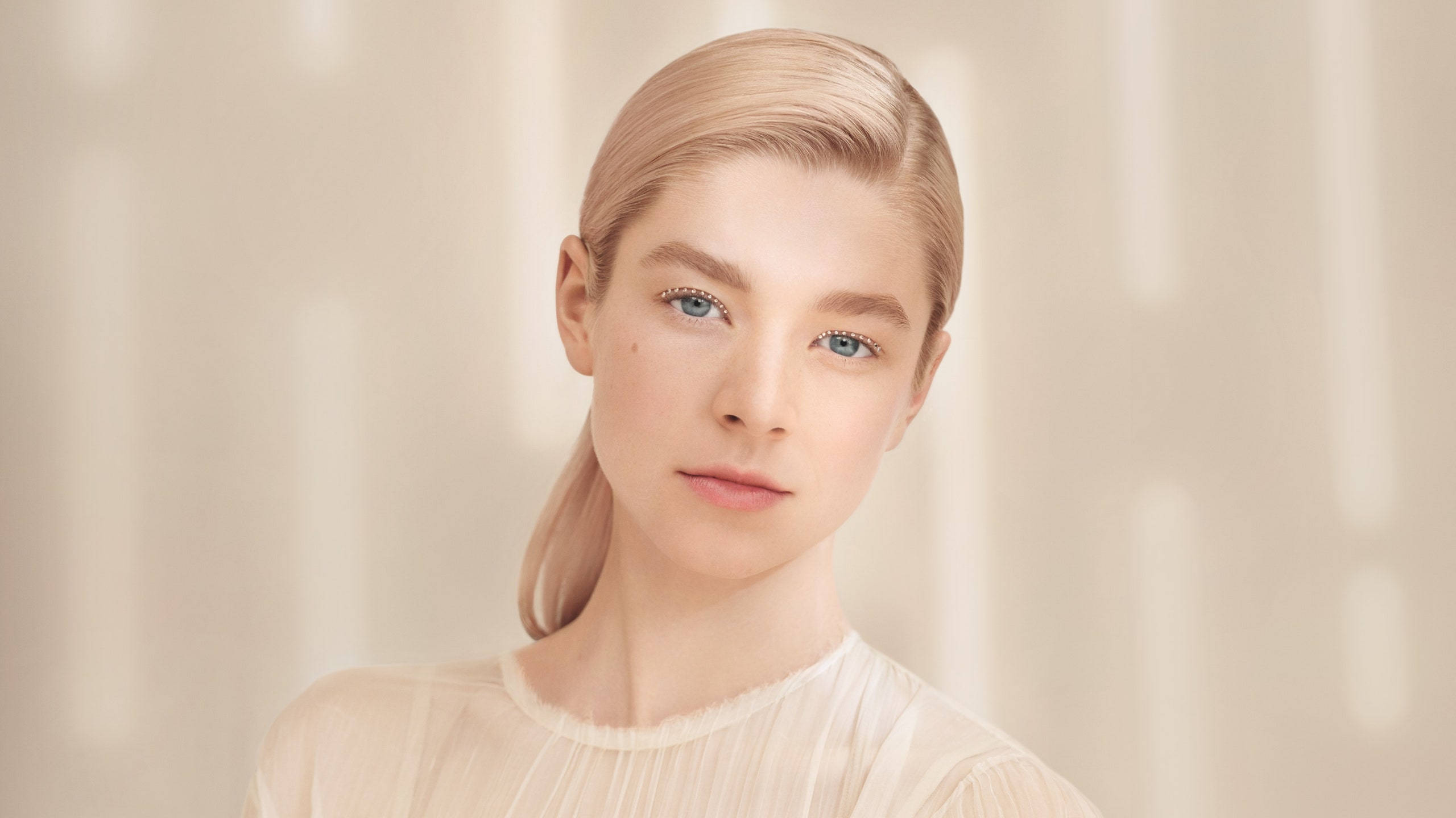Hunter Schafer 桌布 壁紙 — 一位金髮藍眼的年輕女性，身穿透明白色襯衫，在柔和燈光的米色背景下，呈現寧靜的肖像。