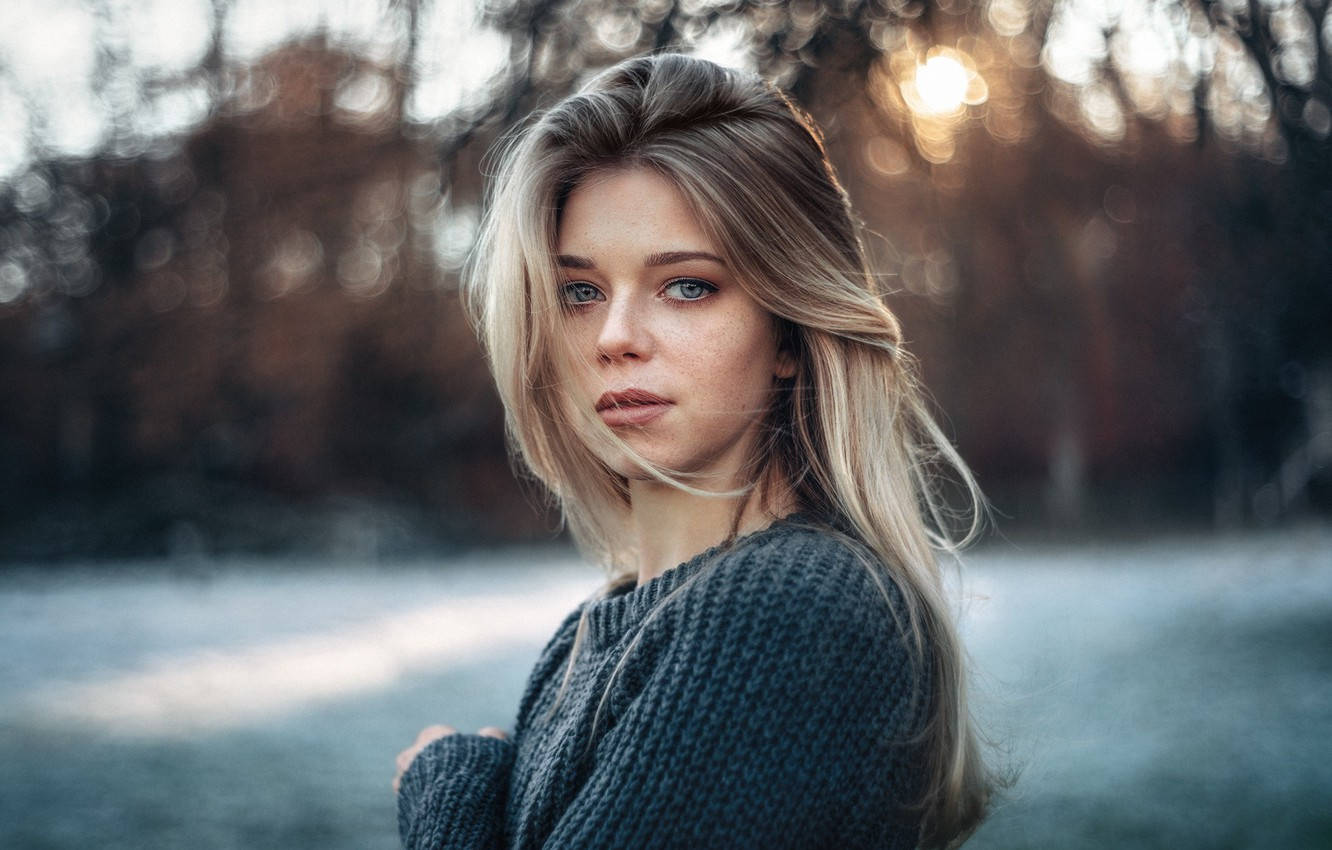 German Girl 桌布 壁紙 — 一位金髮藍眼的年輕女性直視鏡頭，身穿深色毛衣，背景是模糊的秋日森林，金色陽光灑落。