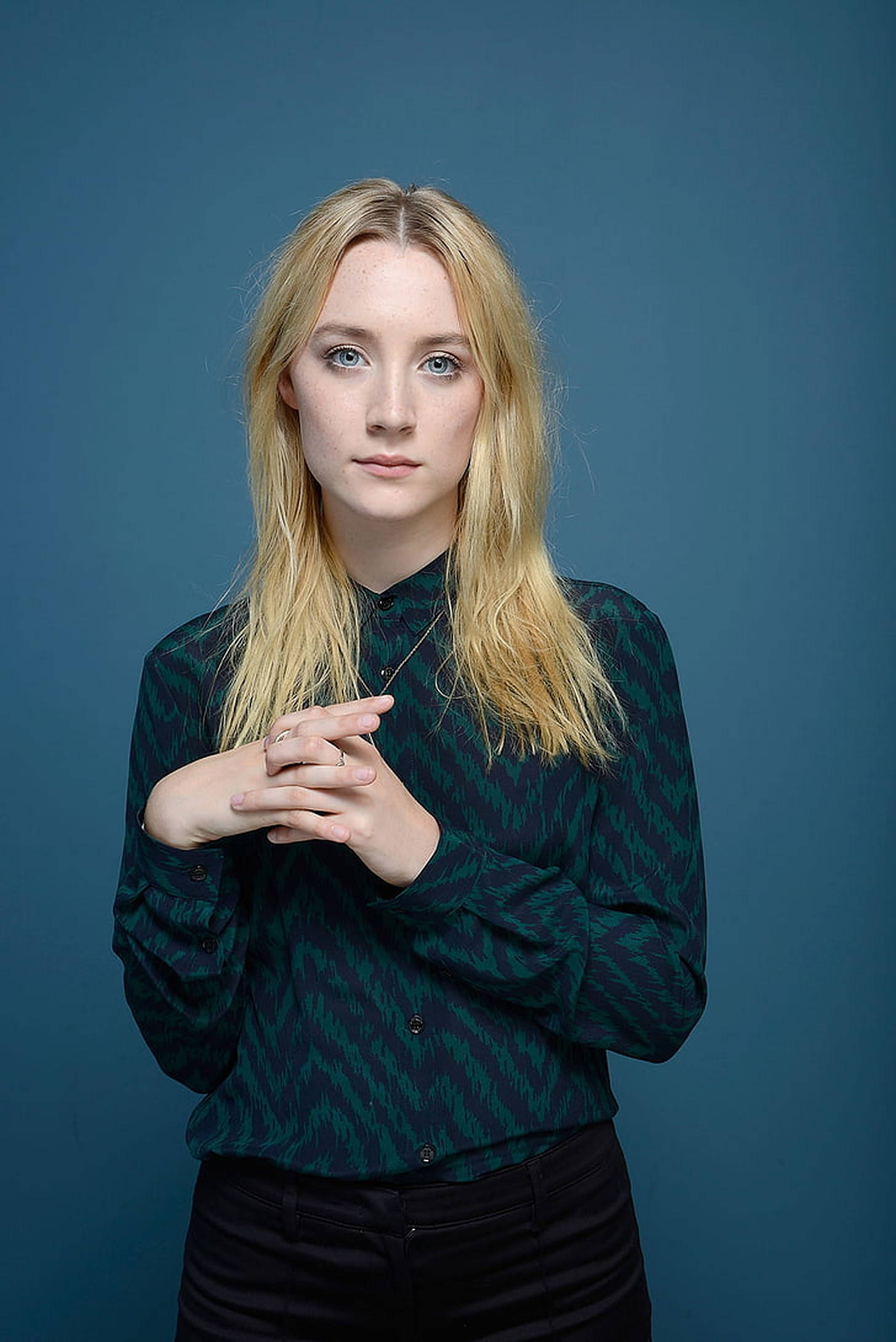 Saoirse Ronan 桌布 壁紙 — 一位金髮藍眼的年輕女性站在純藍色背景前，身穿綠色圖案襯衫和黑色褲子，雙手交握。