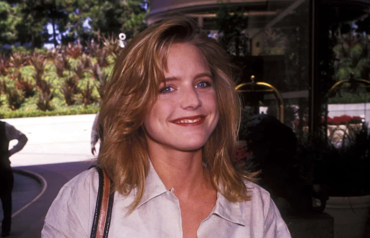 Courtney Thorne Smith 桌布 壁紙 — 一位金髮藍眼的微笑女性，穿著淺色襯衫，在戶外靠近玻璃門，身後有綠植。