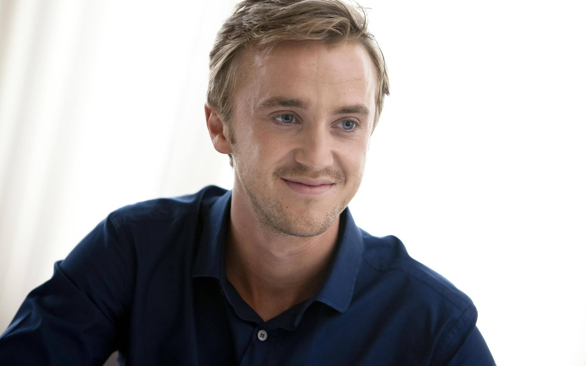 Tom 桌布 壁紙 — 一位金髮藍眼的男士，穿著海軍藍色Polo衫，在明亮的白色背景前溫柔微笑。