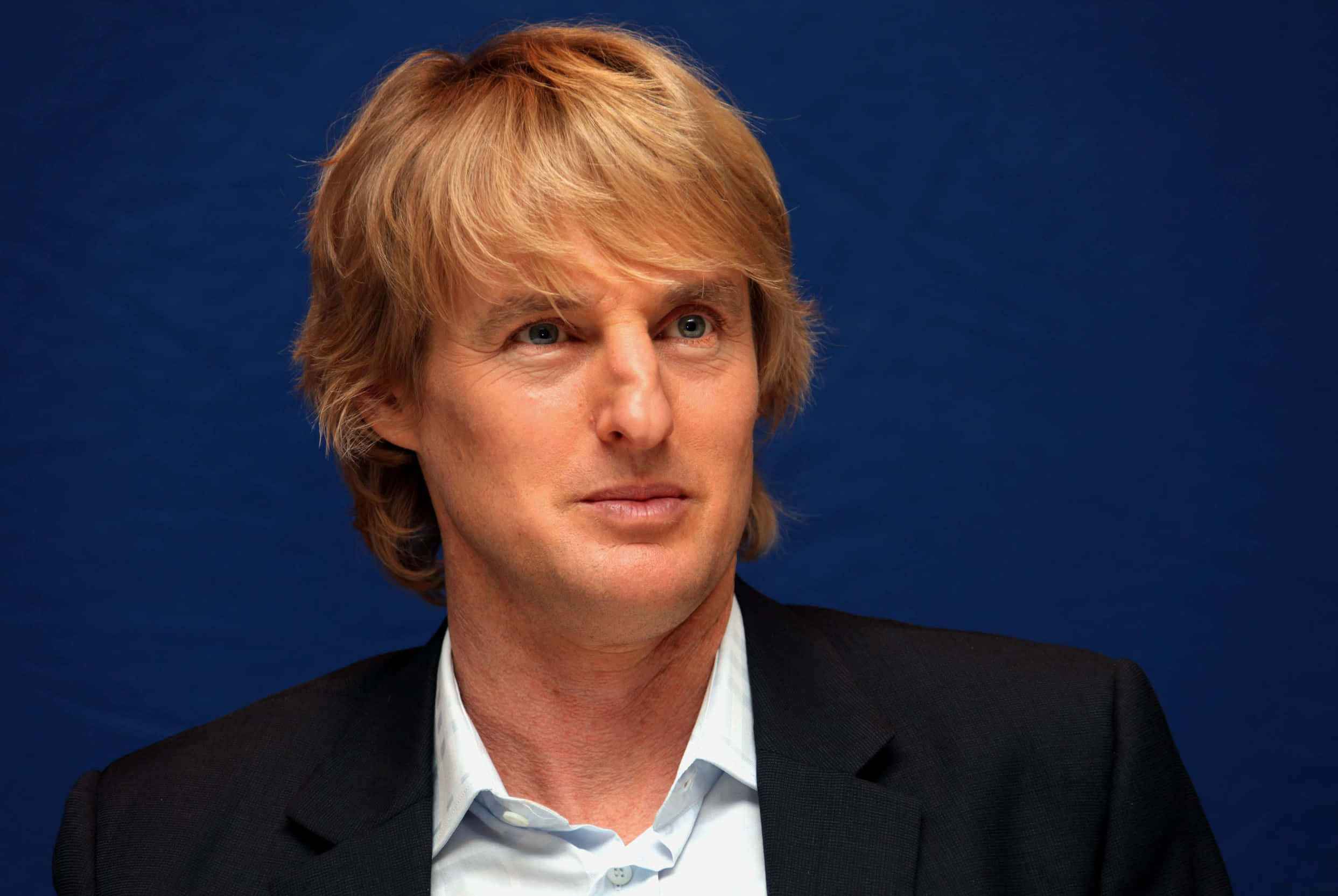 Owen Wilson 桌布 壁紙 — 一位金髮藍眼的男士穿著深色西裝和淺色襯衫，背景為純藍色。