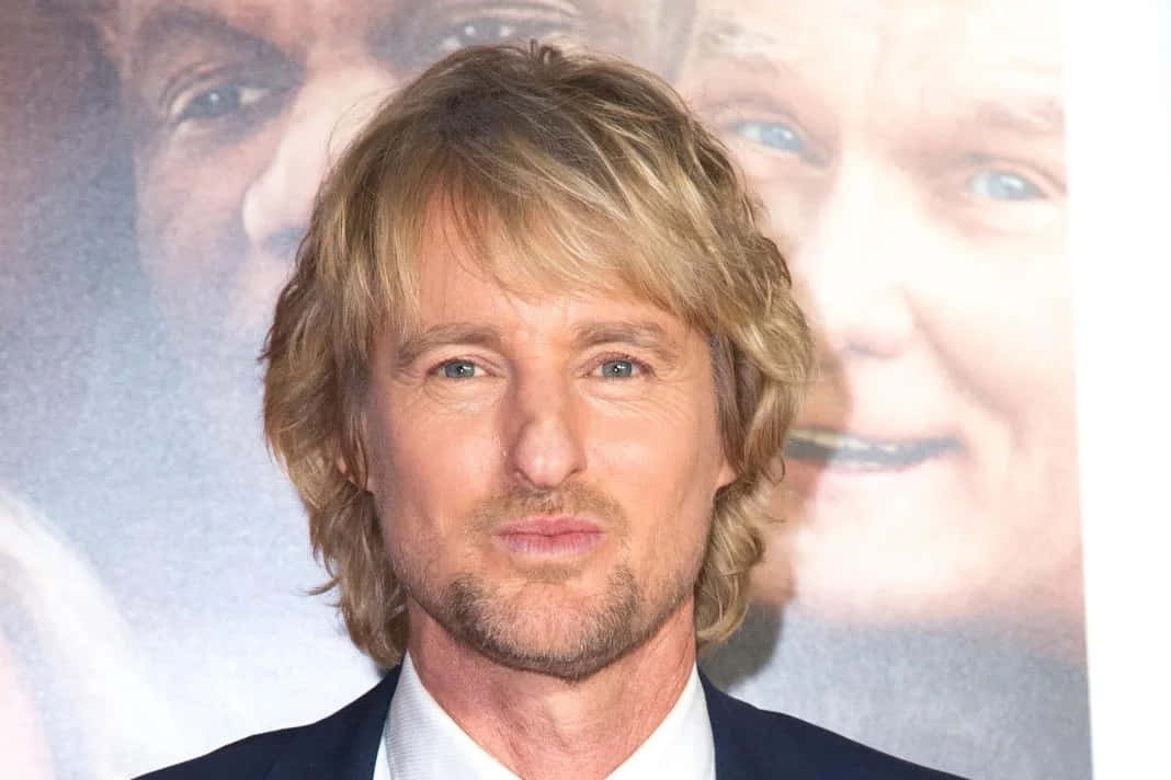 Owen Wilson 桌布 壁紙 — 一位金髮藍眼的男士穿著西裝，站在模糊的電影海報背景前。
