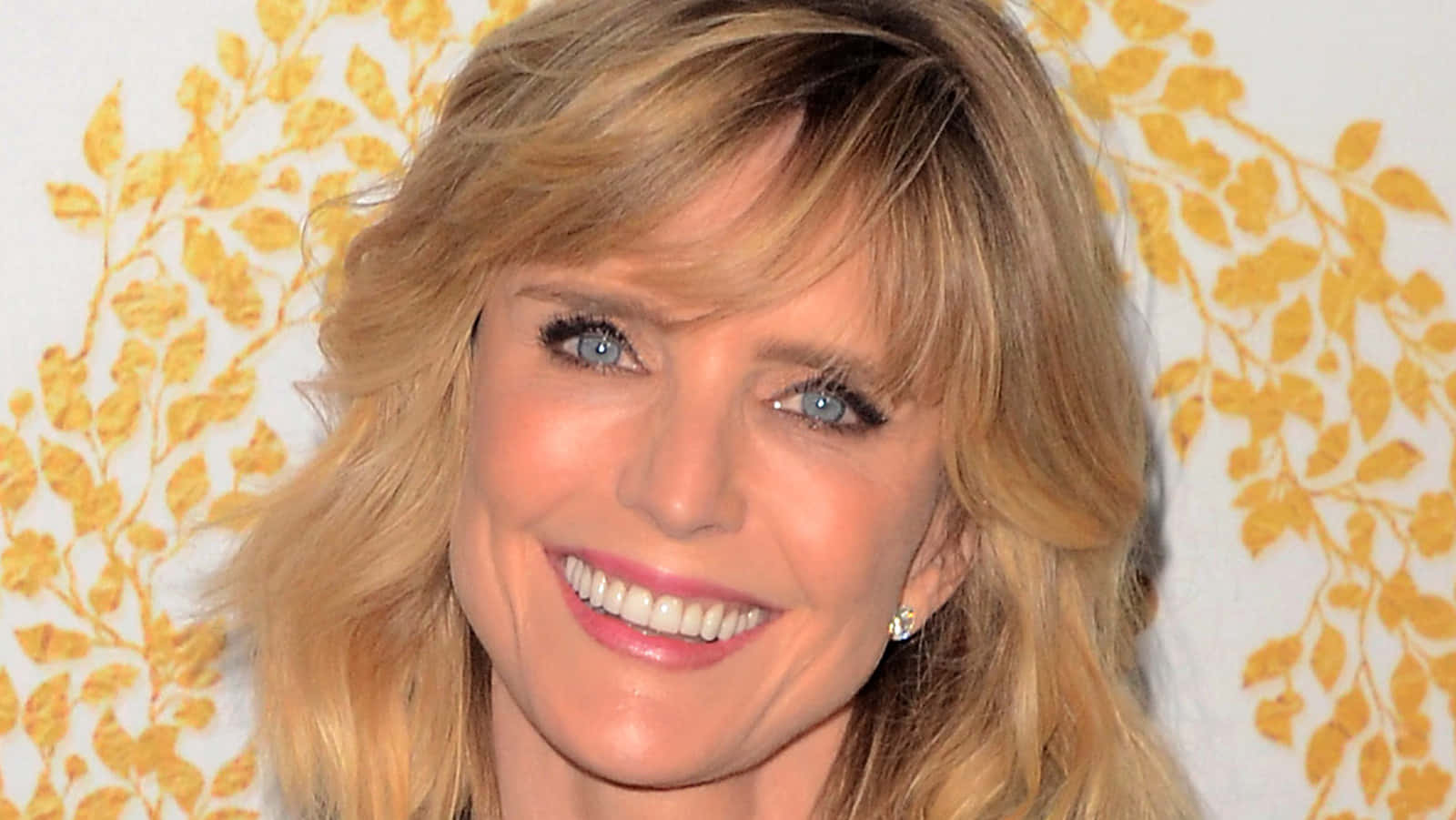 Courtney Thorne Smith 桌布 壁紙 — 一位金髮藍眼的耀眼女性，在黃色花卉壁紙背景前溫暖微笑。