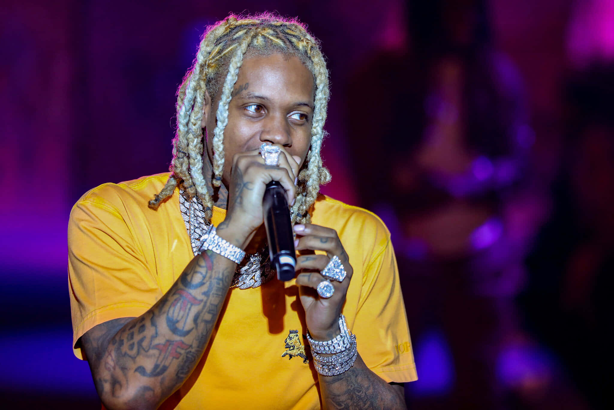 Lil Durk 桌布 壁紙 — 一位金髮辮子、有紋身的饒舌歌手在紫色燈光下穿著黃色襯衫在舞台上表演。
