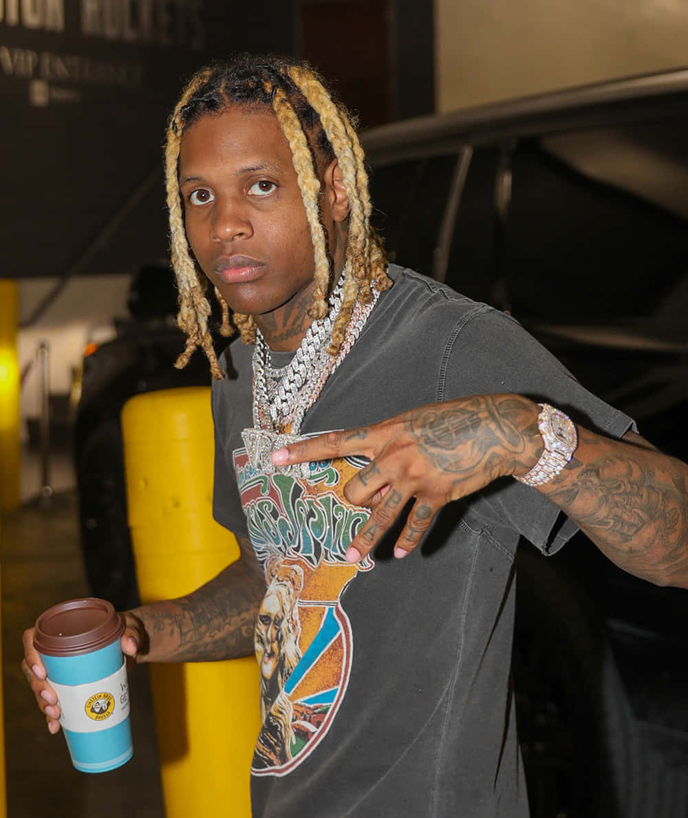 Lil Durk 桌布 壁紙 — 一位金髮辮子刺青男子在昏暗的室內空間自信地拿著咖啡杯擺姿勢。