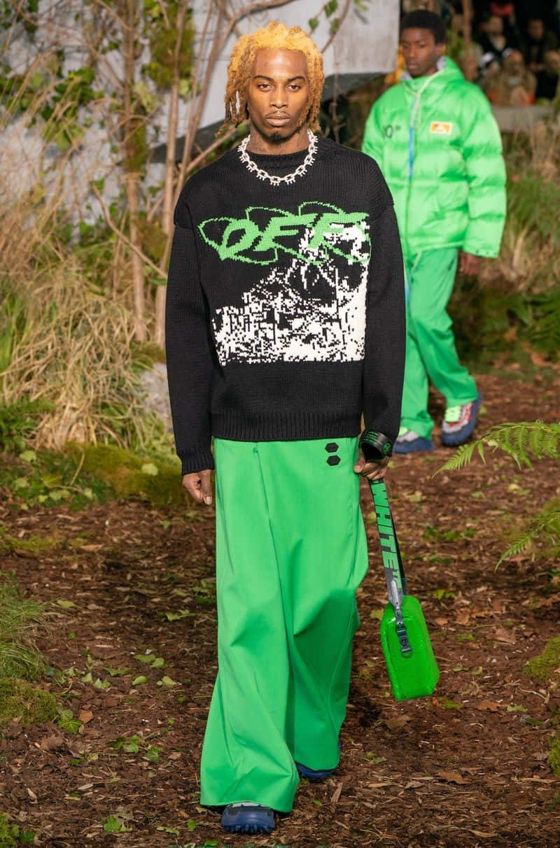 vetements 桌布 壁紙 — 一位金髮辮子模特兒自信地穿著黑色綠色圖案毛衣和亮綠色褲子走在森林小徑上。