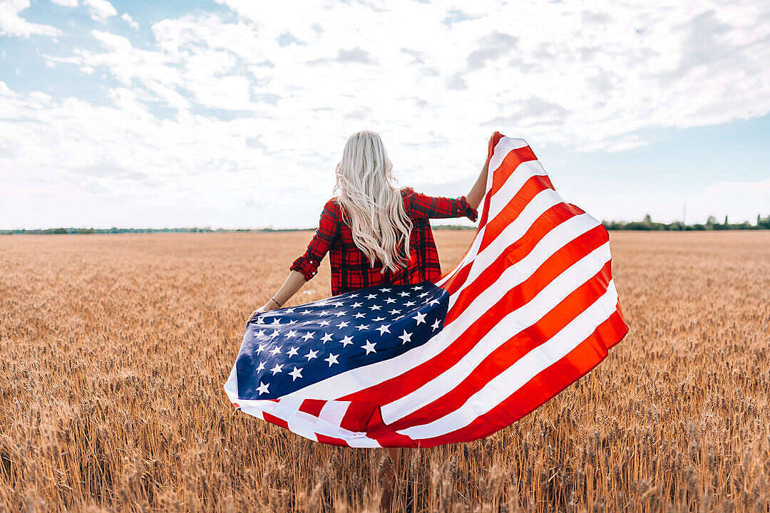 Freedom 桌布 壁紙 — 一位金髮長髮、身穿紅色格紋襯衫的女性，在部分多雲的天空下，於金色麥田中揮舞著美國國旗。