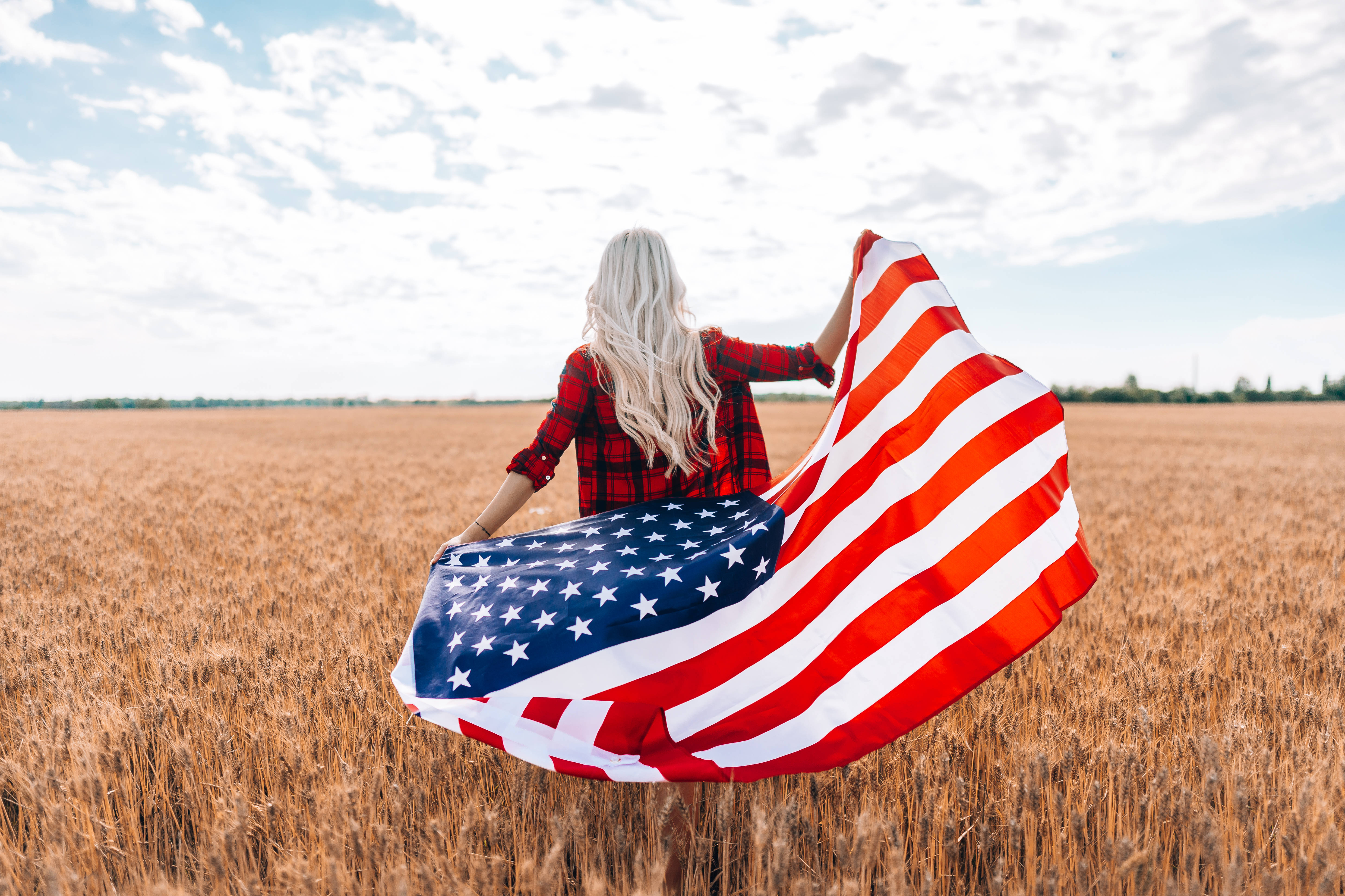 USA flag iPhone 桌布 壁紙 — 一位金髮長髮女性站在金色麥田裡，手持飄揚的美國國旗，背景是部分多雲的天空。