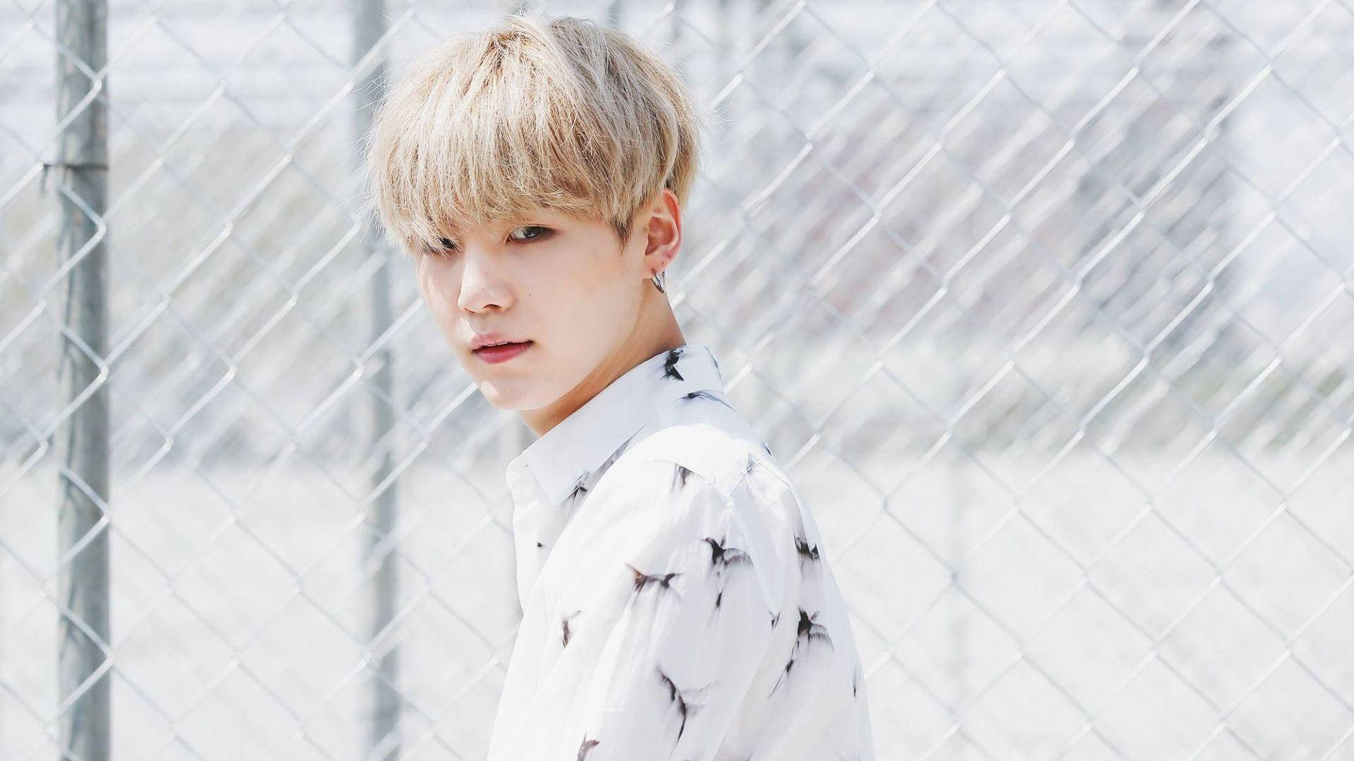 슈가 BTS 桌布 壁紙 — 一位金髮青年穿著星星圖案襯衫，在明亮的日光下站在鐵絲網圍欄前。