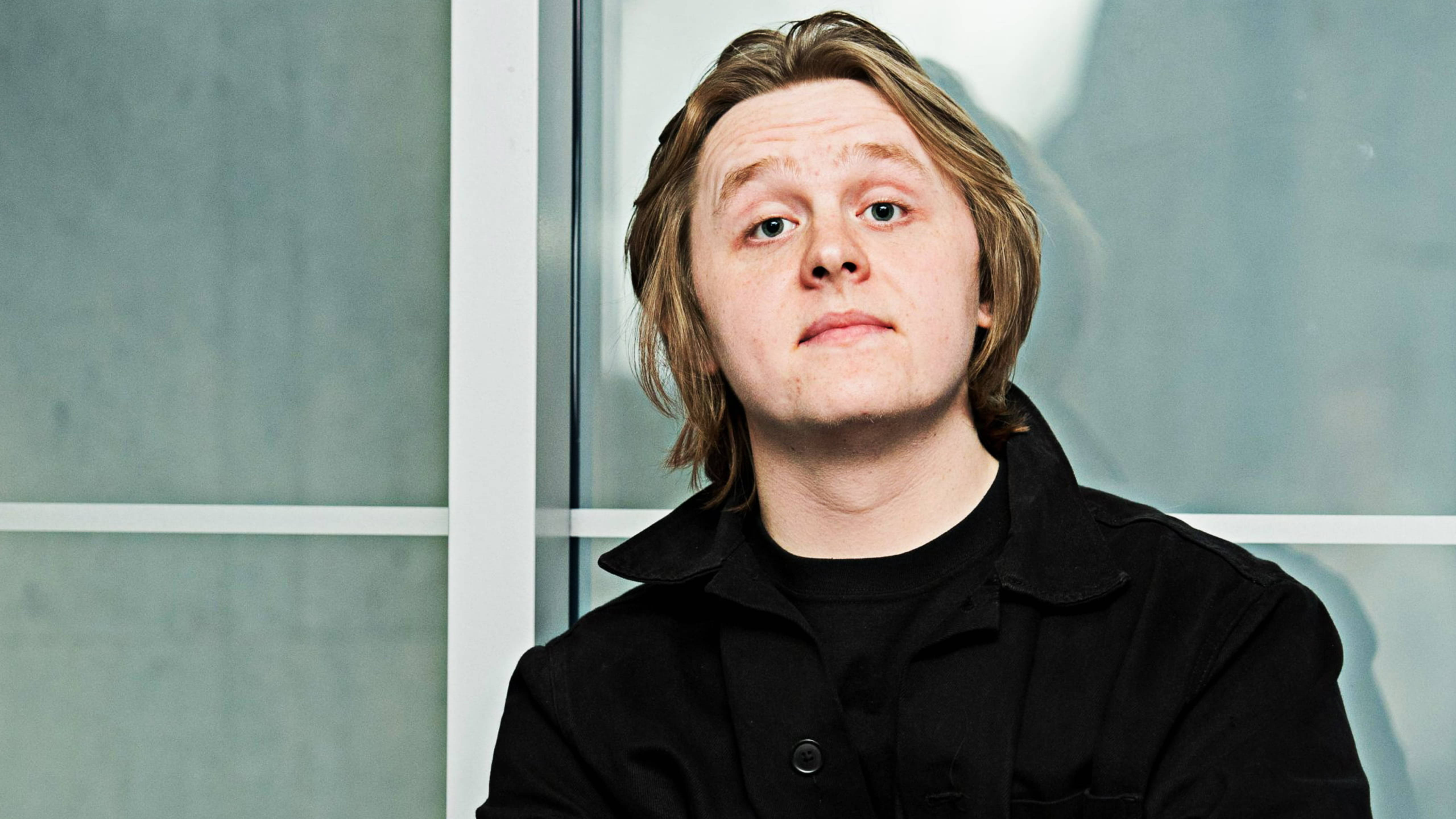 Lewis Capaldi 桌布 壁紙 — 一位金髮青年穿著黑色外套，在現代玻璃面板牆前直視鏡頭。