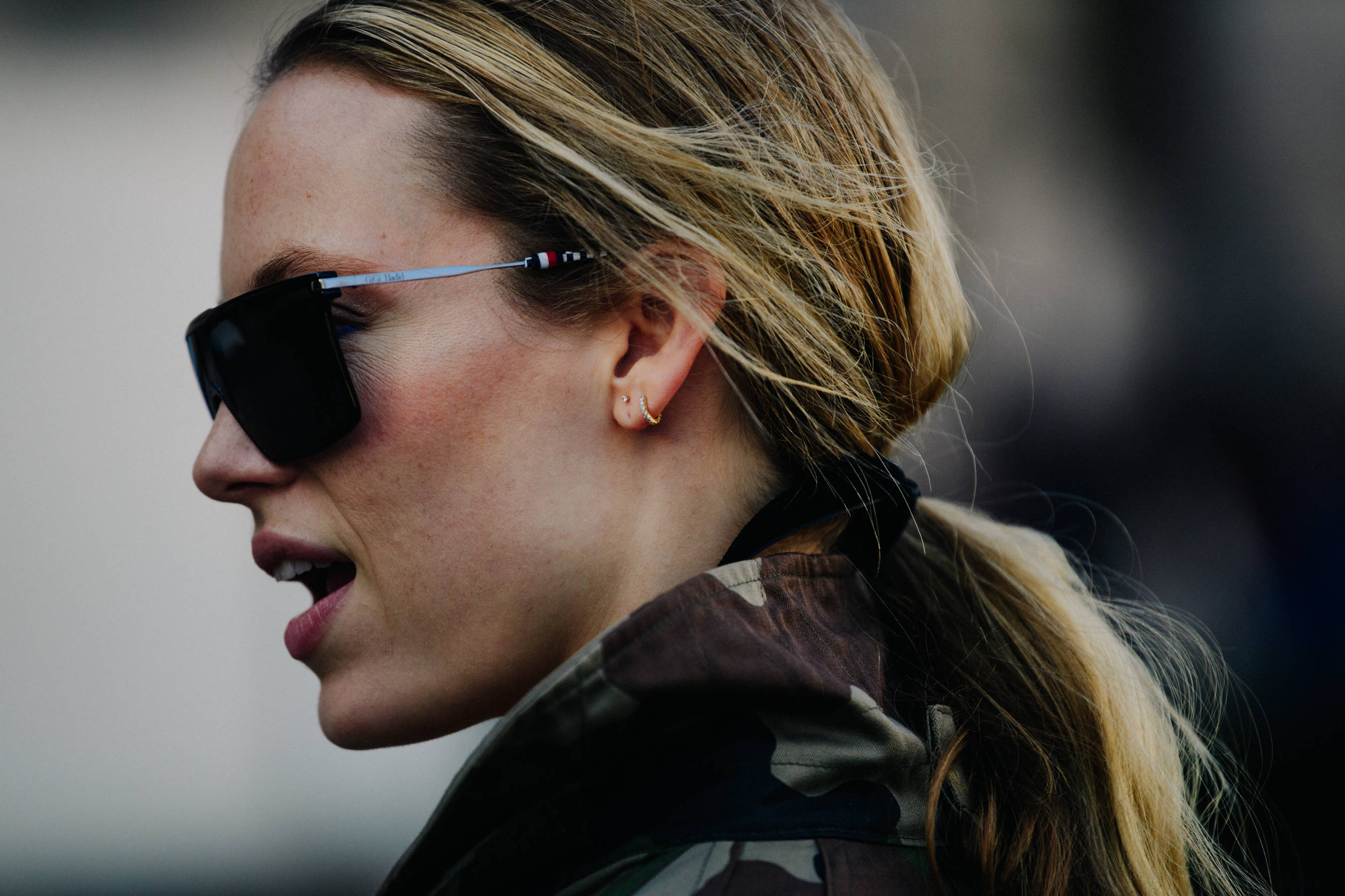 Sunglasses 桌布 壁紙 — 一位金髮馬尾辮的女性戴著黑色太陽眼鏡和迷彩外套，在戶外說話。