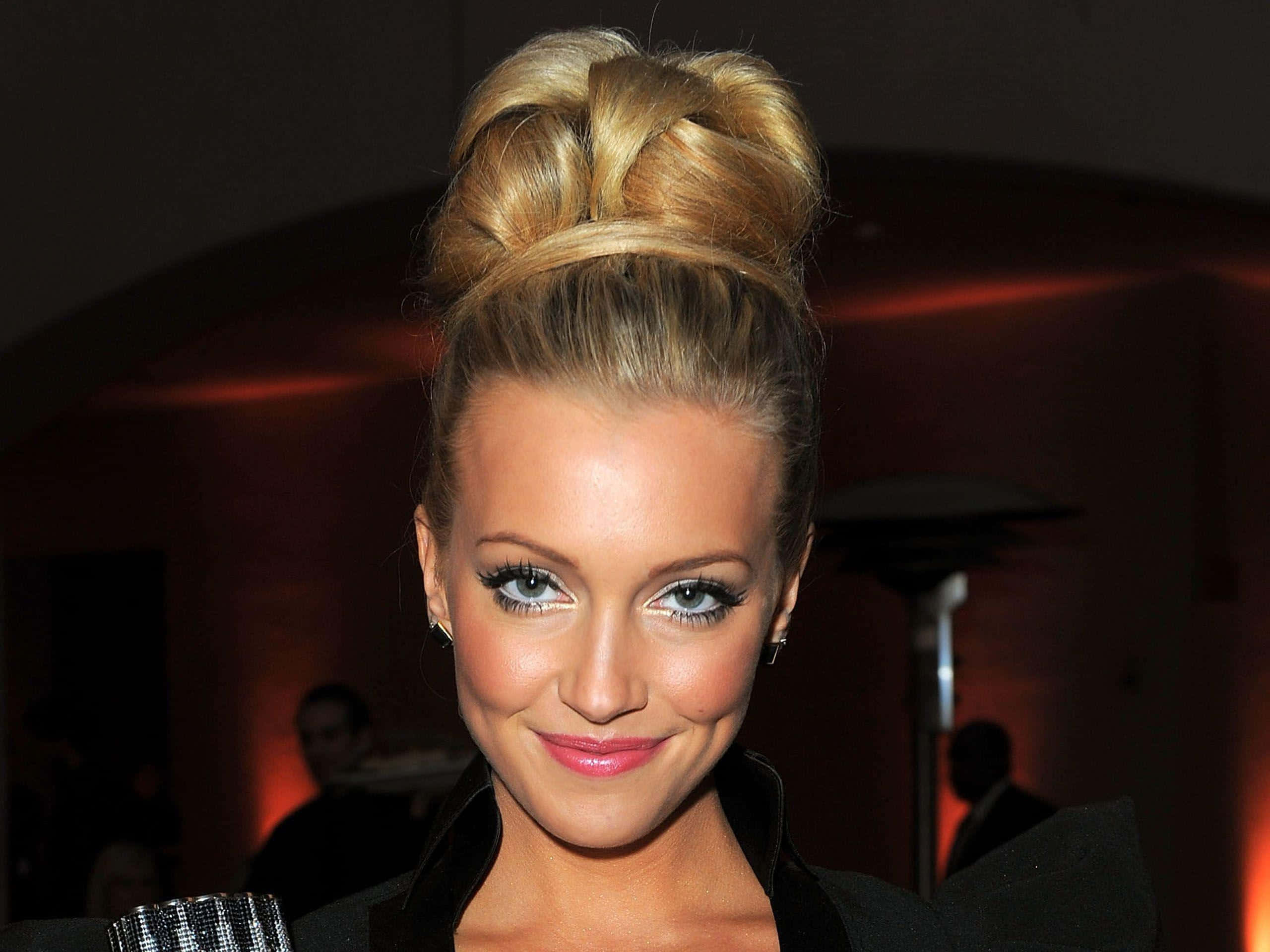 katie cassidy 桌布 壁紙 — 一位金髮高髻的女性在昏暗的室內環境中對著鏡頭溫暖地微笑。
