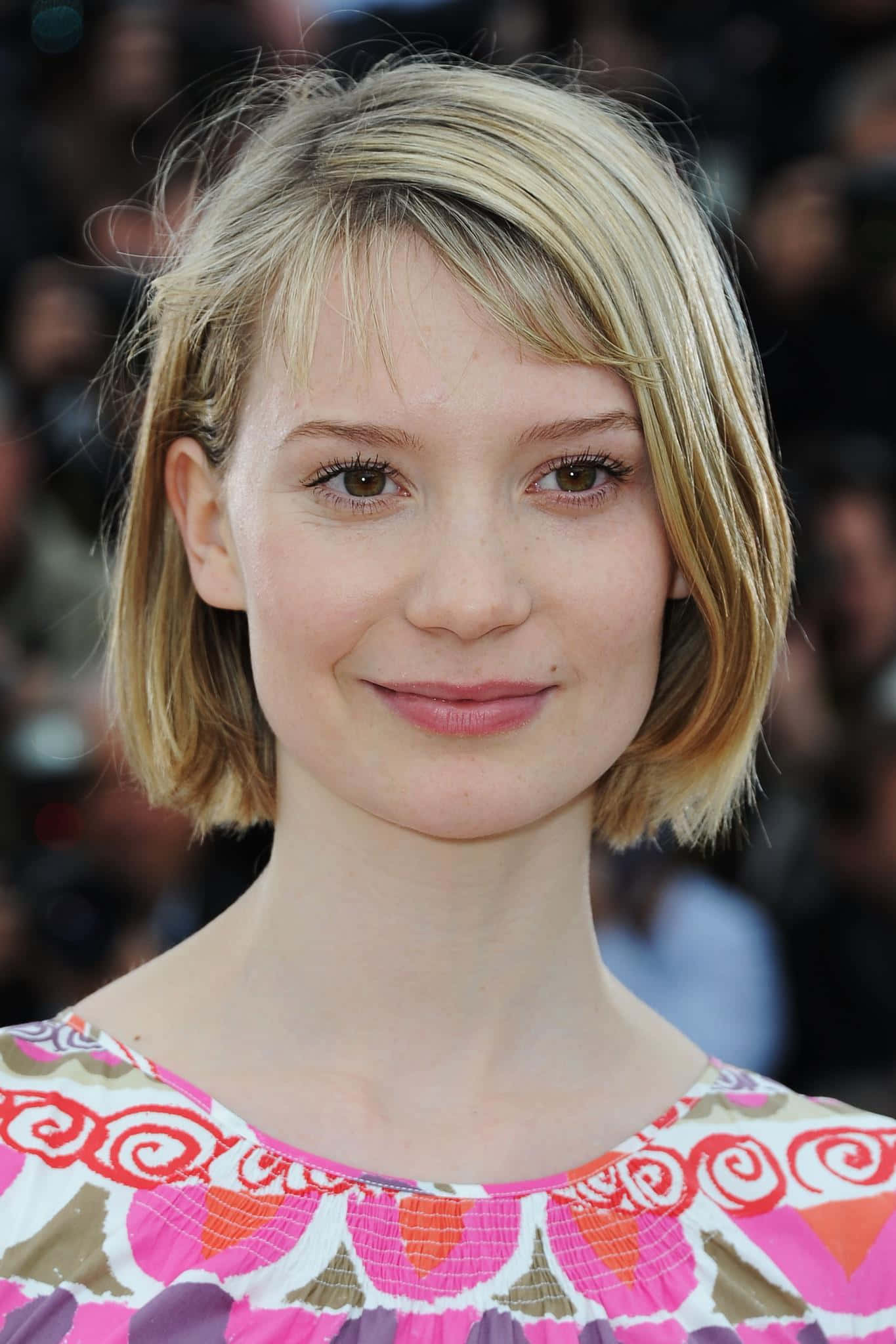 Mia Wasikowska 桌布 壁紙 — 一位金髮鮑伯頭女性的特寫肖像，帶著溫柔的笑容，身穿色彩繽紛的圖案上衣。