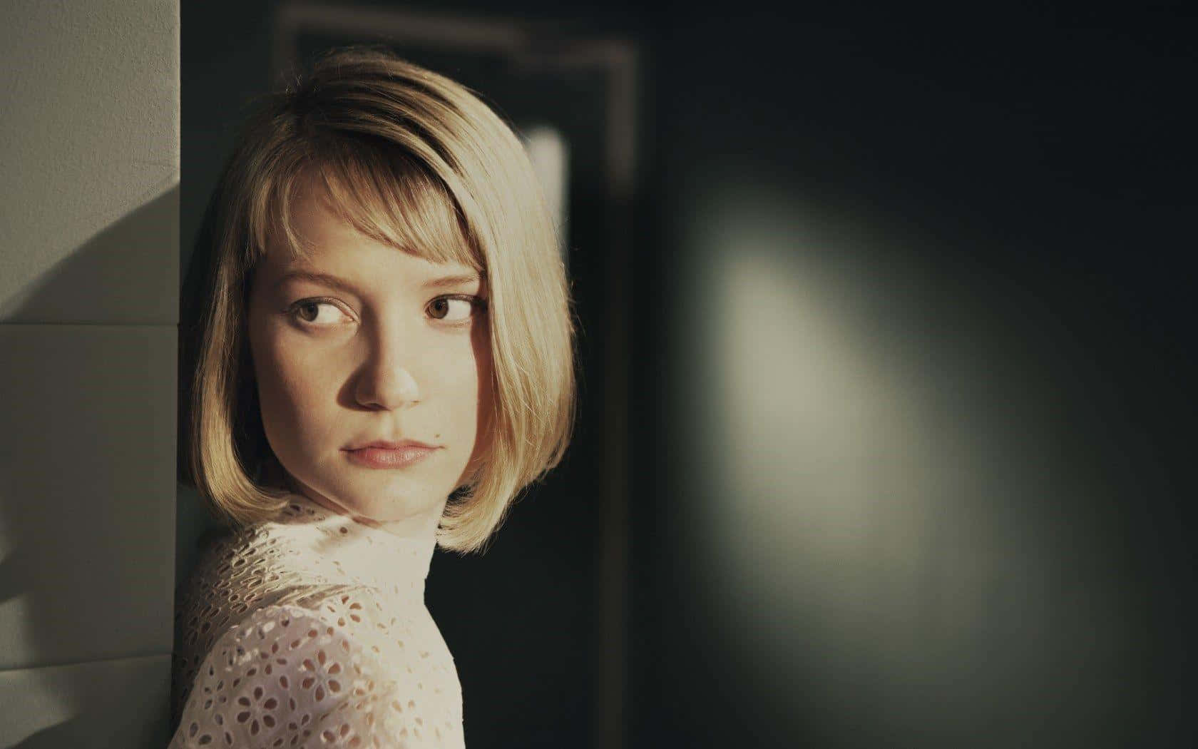 Mia Wasikowska 桌布 壁紙 — 一位金髮鮑伯頭的年輕女性在昏暗的房間中，從牆後探出頭來，帶有戲劇性的側光。