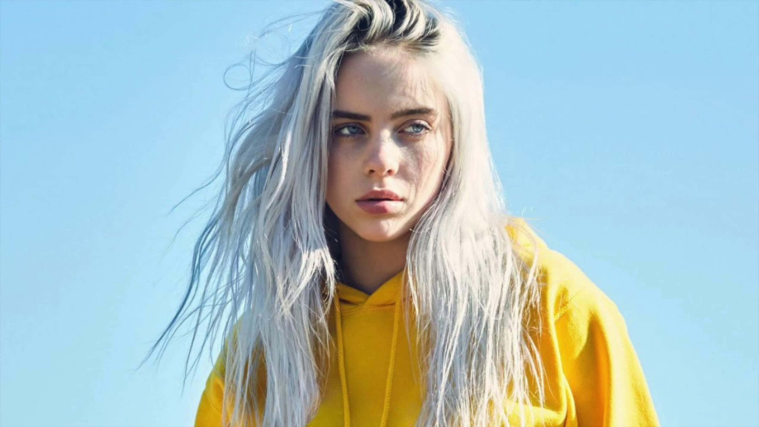 Billie Eilish Laptop 桌布 壁紙 — 一位銀色頭髮、穿著黃色連帽衫的年輕女子，在清澈的藍天下強烈地凝視著。