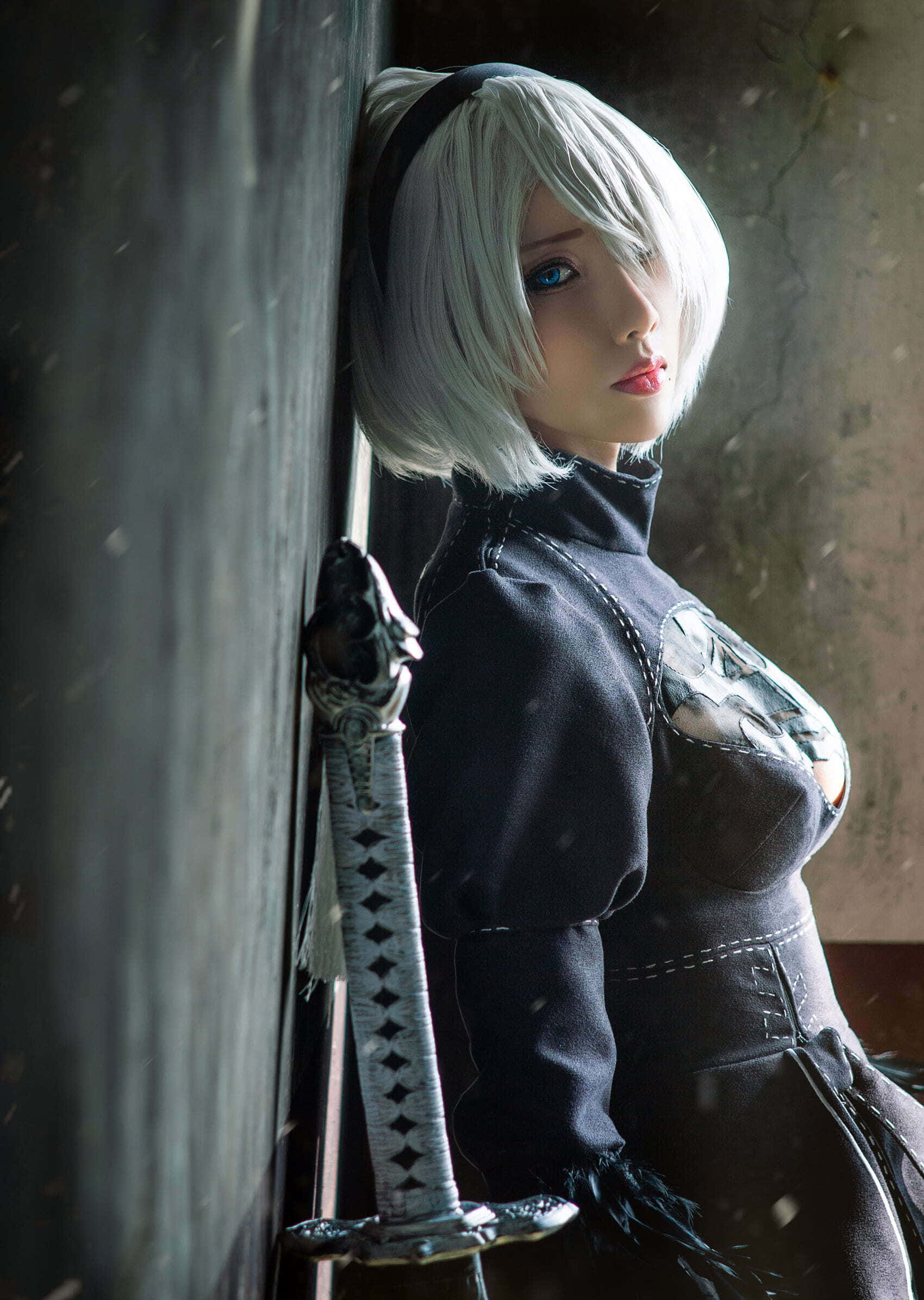 Gaming Cosplay 桌布 壁紙 — 一位銀色鮑伯頭、藍眼睛的角色扮演者靠在牆上，在昏暗的雪景中手持一把精緻的劍。