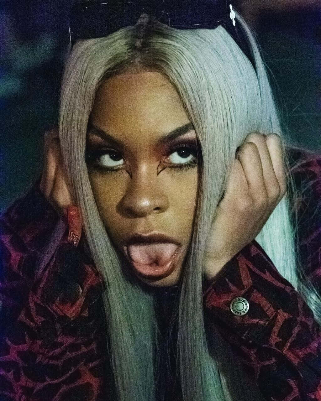Rico Nasty 桌布 壁紙 — 一位銀髮且妝容大膽的人，在昏暗光線下穿著圖案外套伸出舌頭。