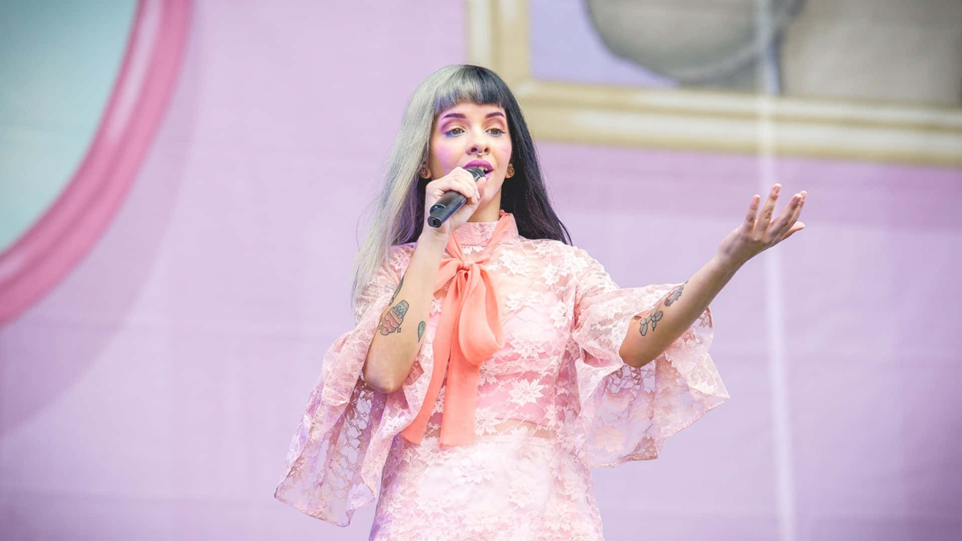 Melanie Martinez 桌布 壁紙 — 一位銀髮刺青女歌手身穿粉紅色蕾絲服裝在舞台上手持麥克風表演。