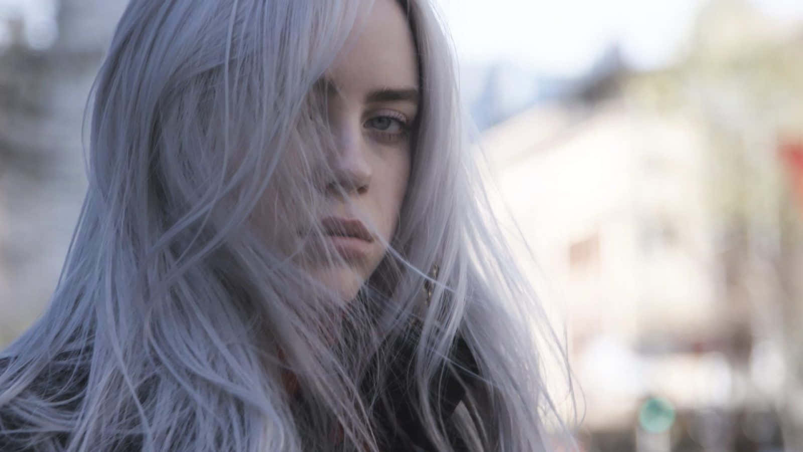 Billie Eilish Laptop 桌布 壁紙 — 一位銀髮女子眼神專注地向前望，風吹動髮絲掠過她的臉龐，背景是模糊的城市景觀。