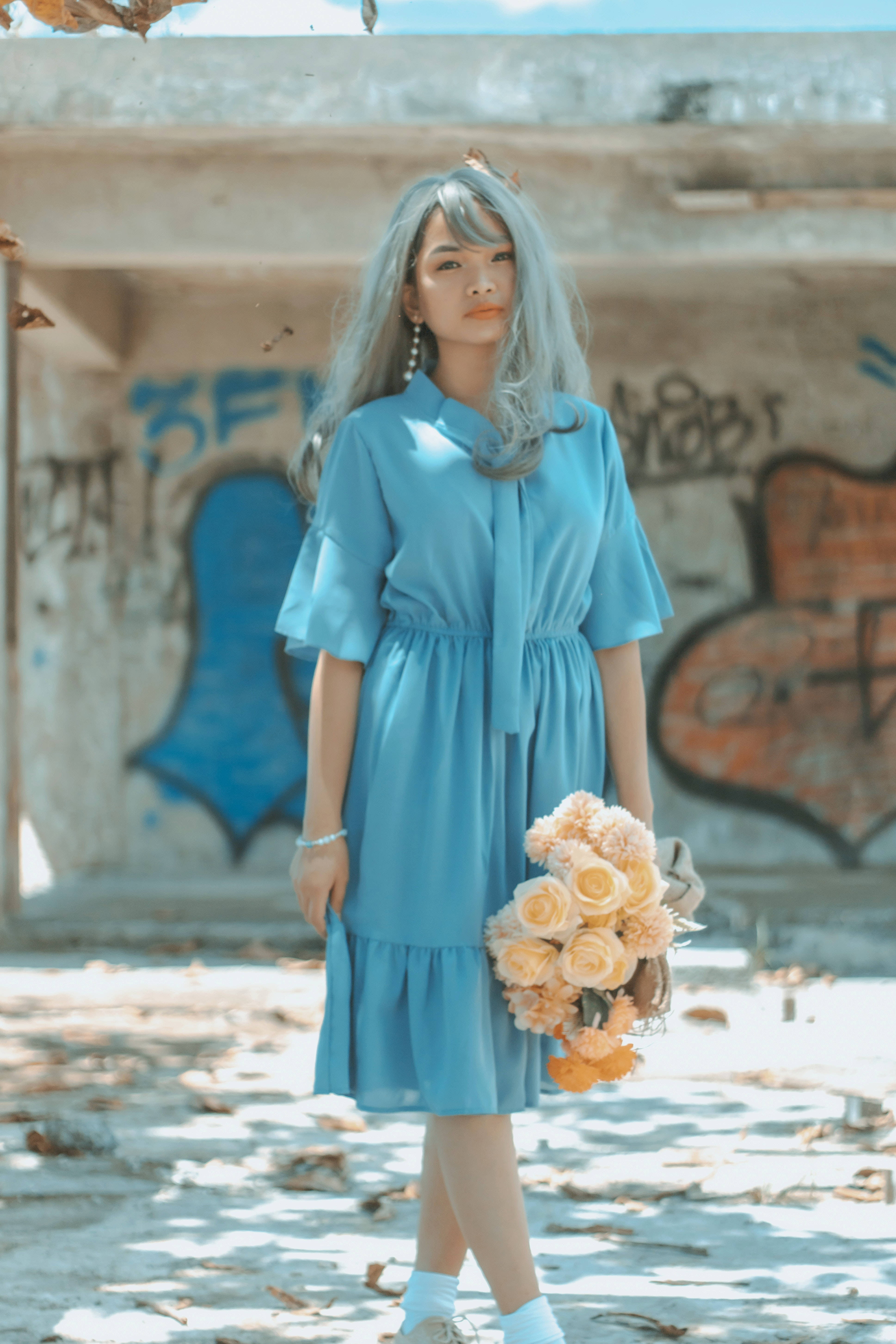 Cute Blue 桌布 壁紙 — 一位銀髮年輕女子身穿藍色連衣裙，在明亮的陽光下，手持一束蜜桃玫瑰，背景是塗鴉覆蓋的城市廢墟。