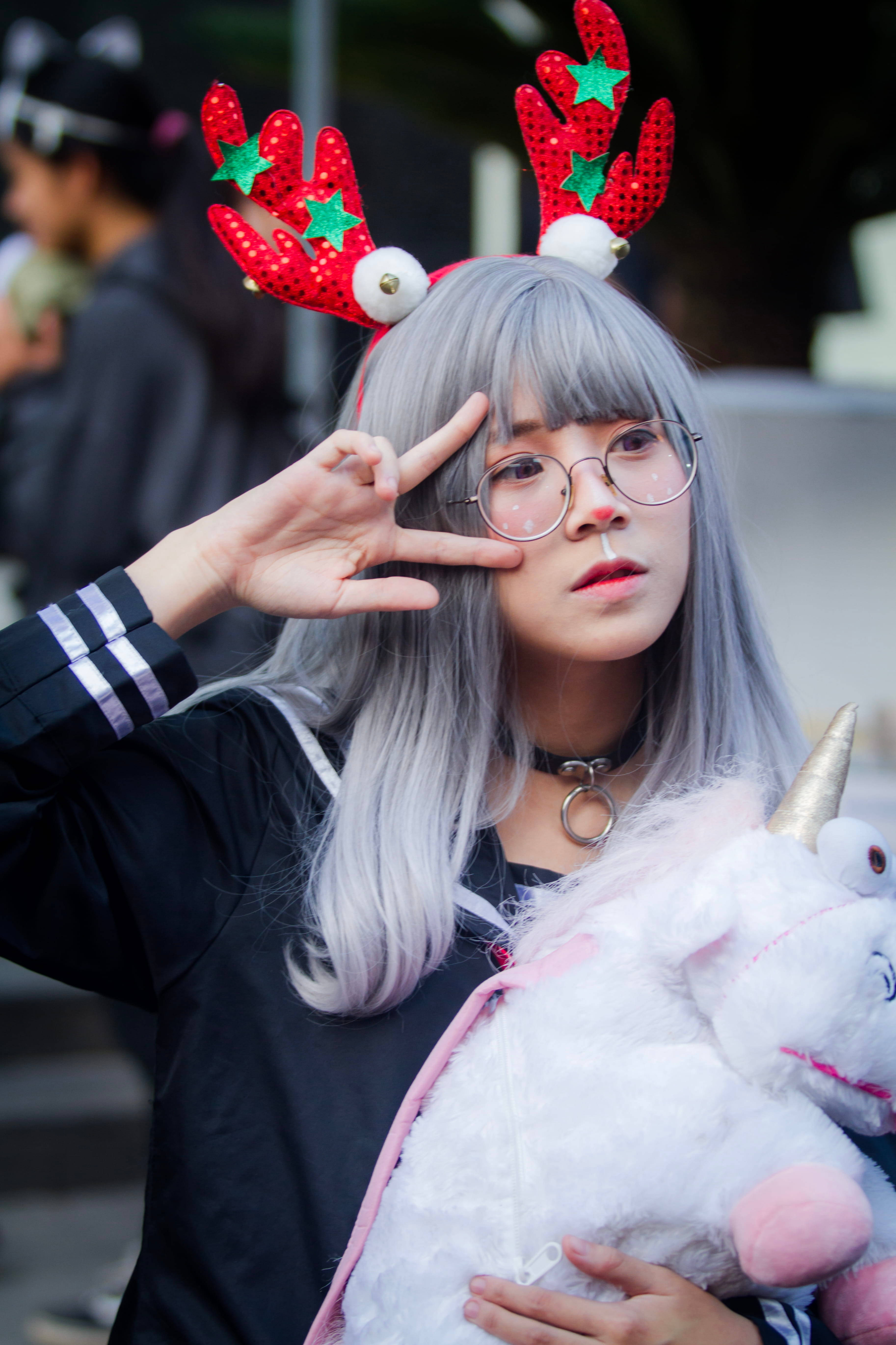 東京 桌布 壁紙 — 一位銀髮戴著馴鹿角的年輕女子，抱著獨角獸毛絨玩具，比出和平手勢。