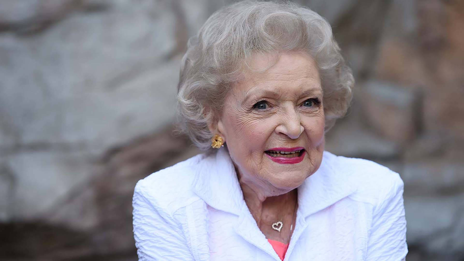 Betty White 桌布 壁紙 — 一位銀髮紅唇的老婦人，在模糊的石牆背景下溫暖地微笑。