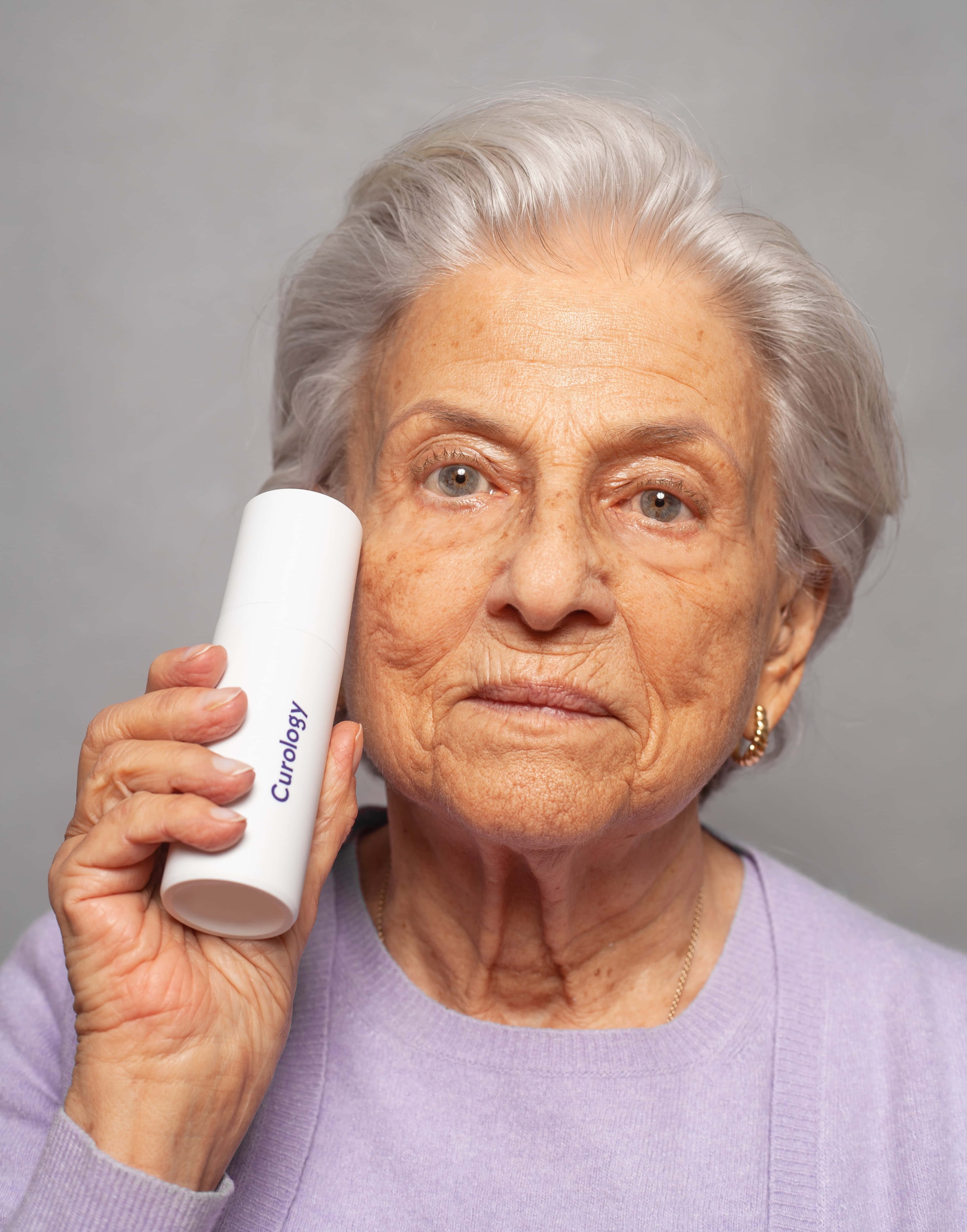 Mature 桌布 壁紙 — 一位銀髮老婦人將白色「Curology」管狀容器貼在耳邊，彷彿在傾聽，背景為純灰色。