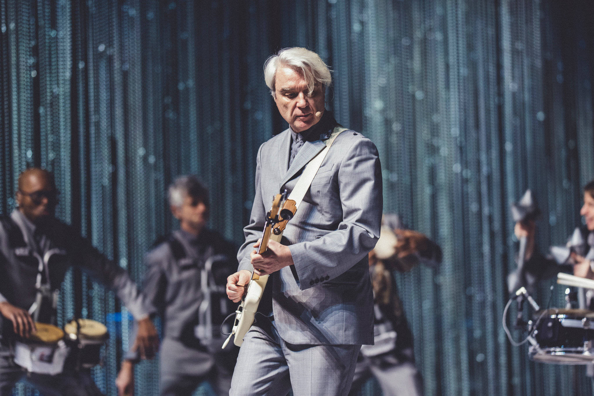 band 桌布 壁紙 — 一位銀髮音樂家身穿灰色西裝，在閃爍藍色帷幕背景的舞台上彈奏白色電吉他。