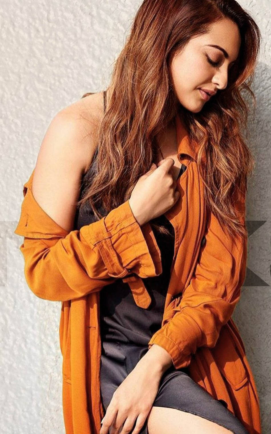 Sonakshi Sinha 桌布 壁紙 — 一位長捲髮的女性穿著橙色開襟襯衫，靠在有紋理的白色牆壁上，低頭露出柔和的表情。