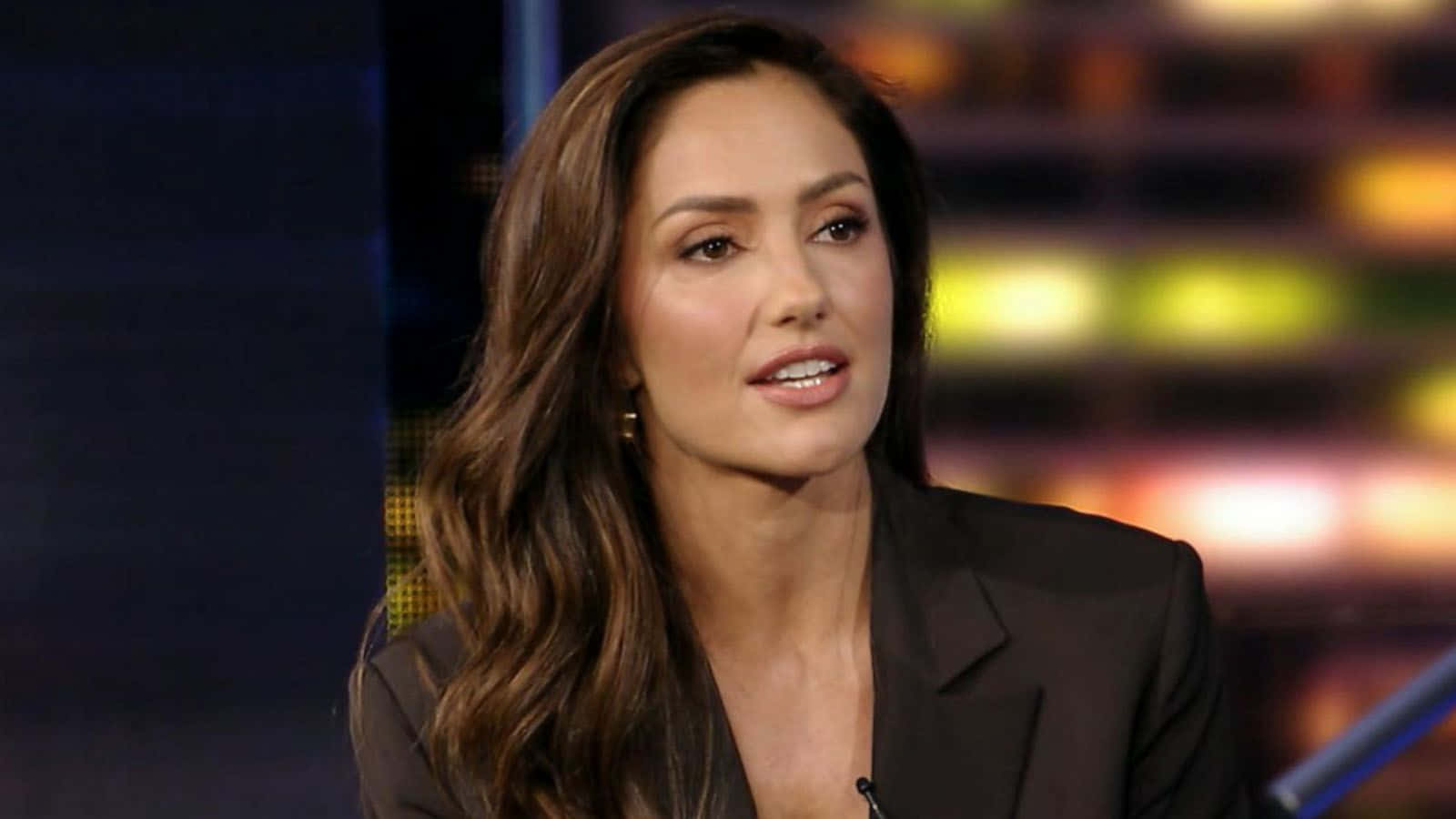 minka kelly 桌布 壁紙 — 一位長棕色頭髮、身穿黑色西裝外套的女性在模糊的彩色背景前說話。
