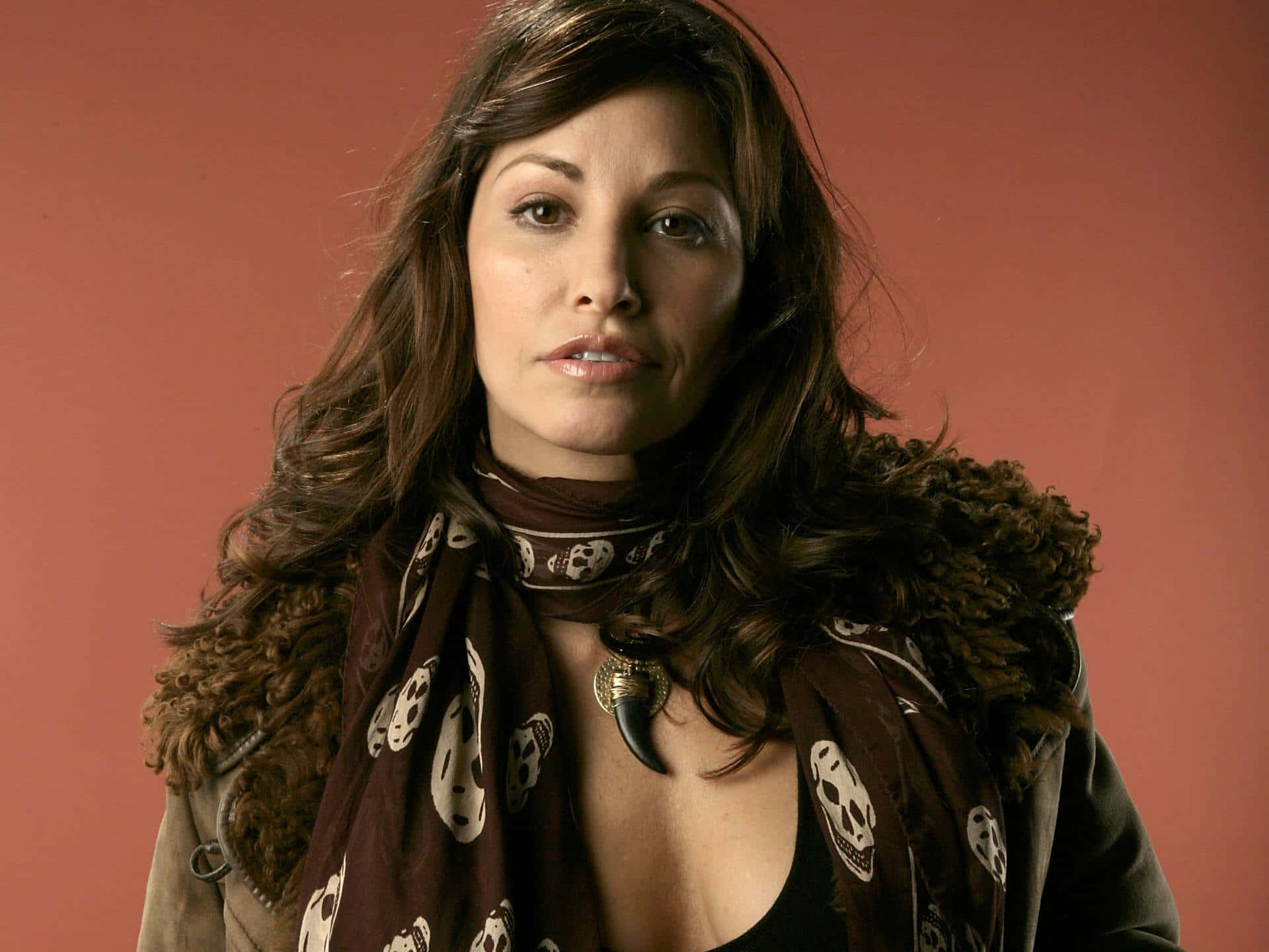 Gina Gershon 桌布 壁紙 — 一位長棕色頭髮的女性，在純色陶土背景前，戴著骷髏圖案圍巾和毛邊夾克。