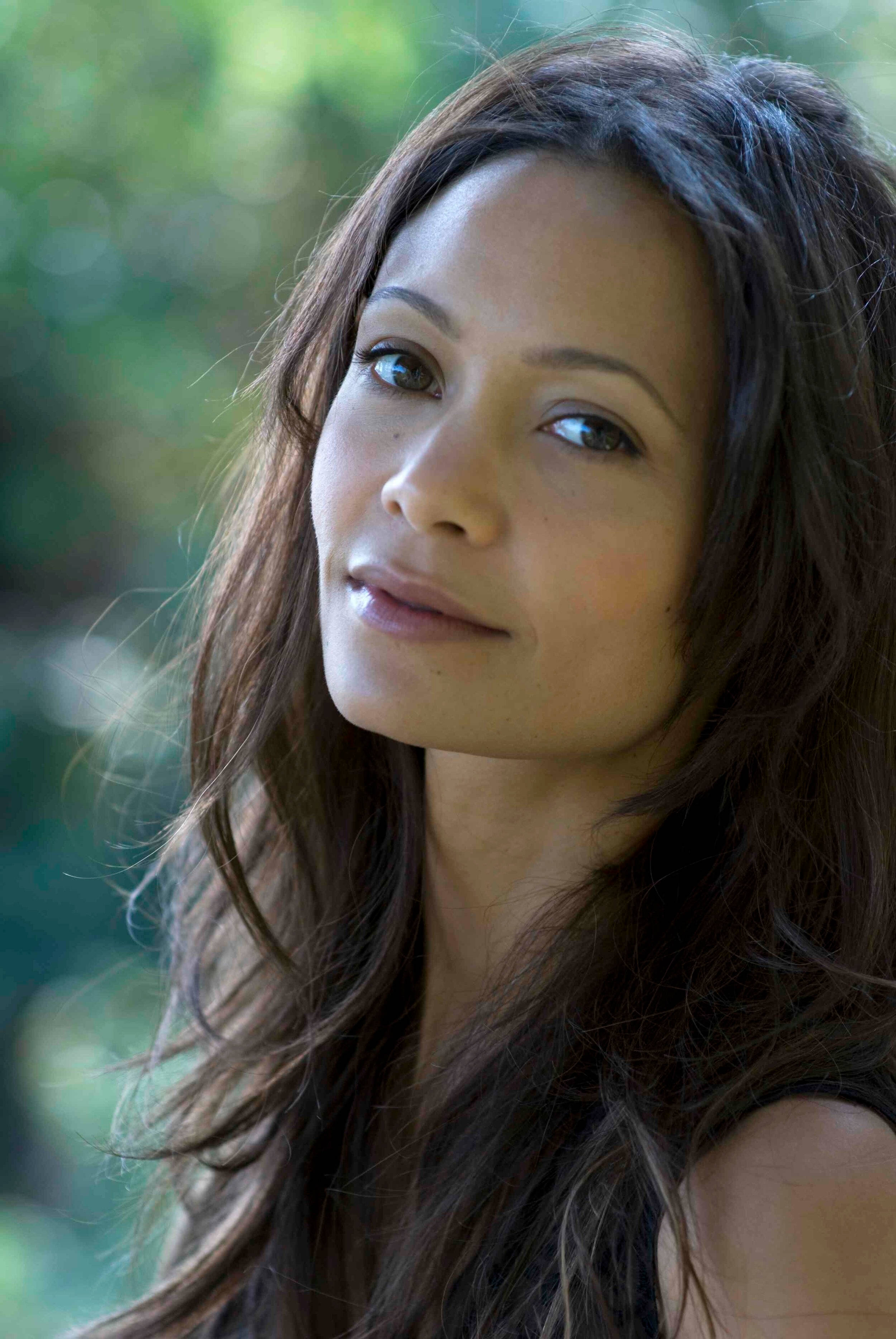 Actress HD 桌布 壁紙 — 一位長棕色頭髮的女性在模糊的綠色樹葉背景下溫柔地凝視著鏡頭。