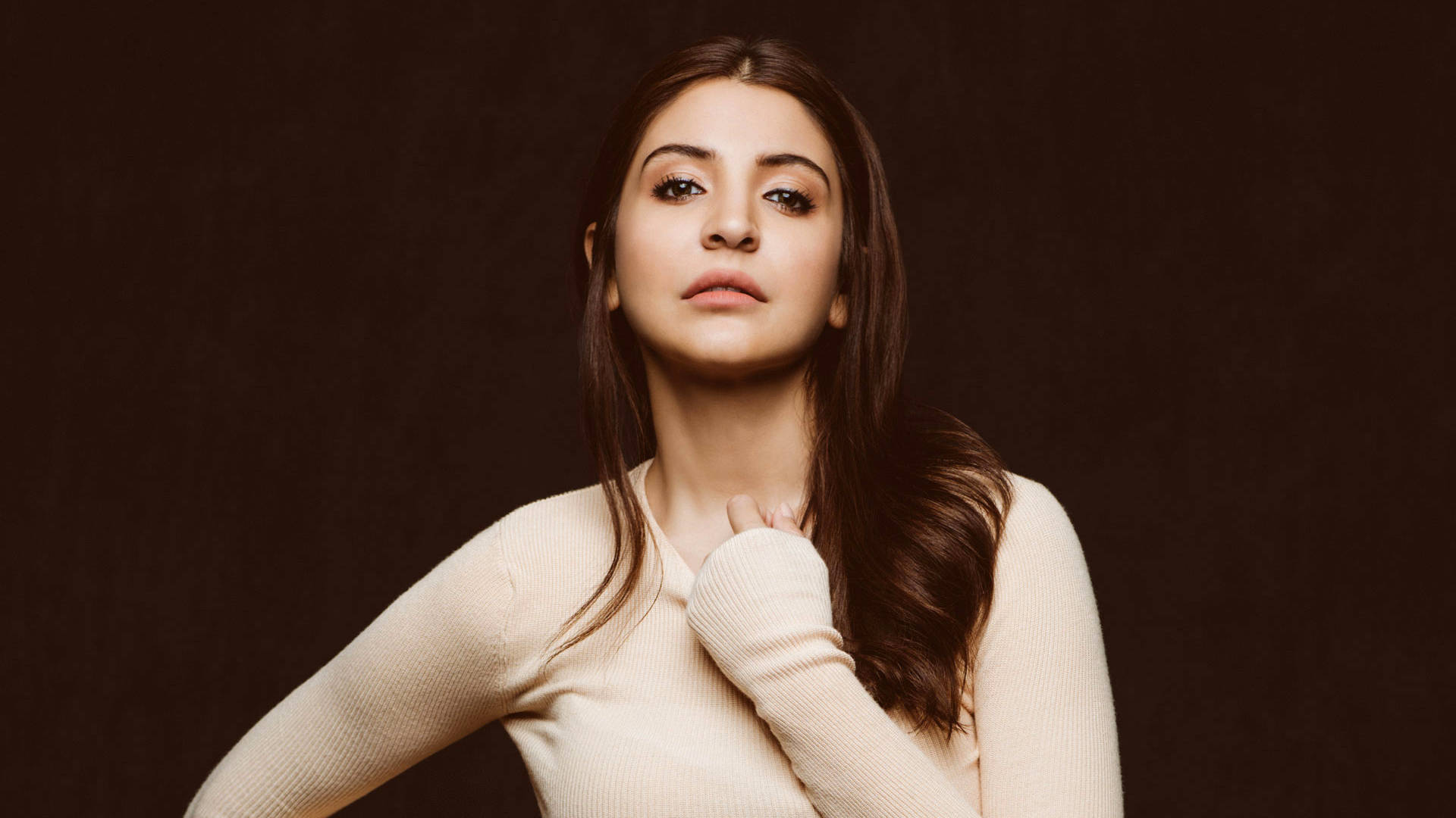 Anushka Sharma 桌布 壁紙 — 一位長棕色頭髮的女性在深棕色背景前穿著奶油色羅紋毛衣，在攝影棚拍攝中向上凝視。