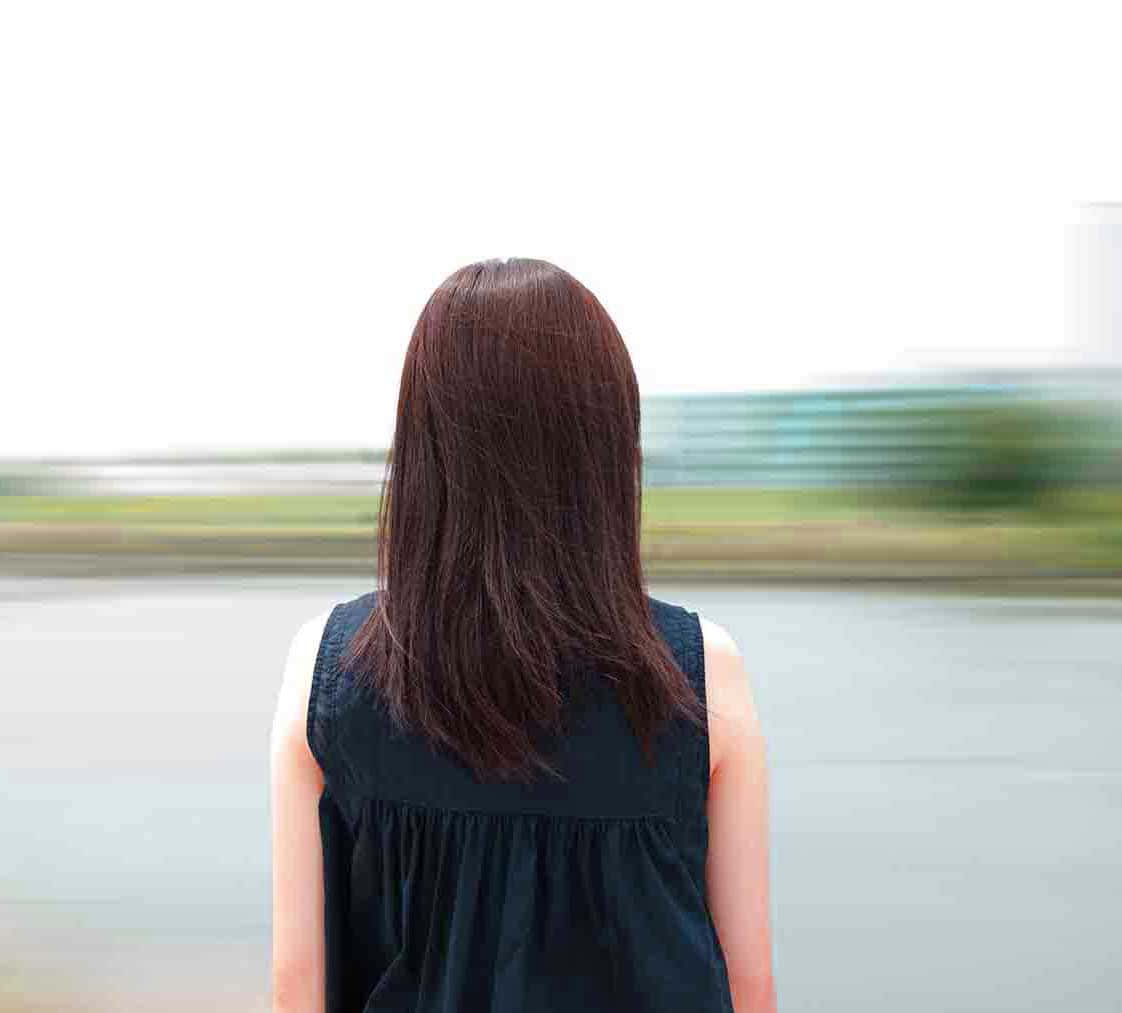 Depressing Profile 桌布 壁紙 — 一位長棕色頭髮的女性背對鏡頭站立，在柔和日光下凝視著模糊的路邊景色。