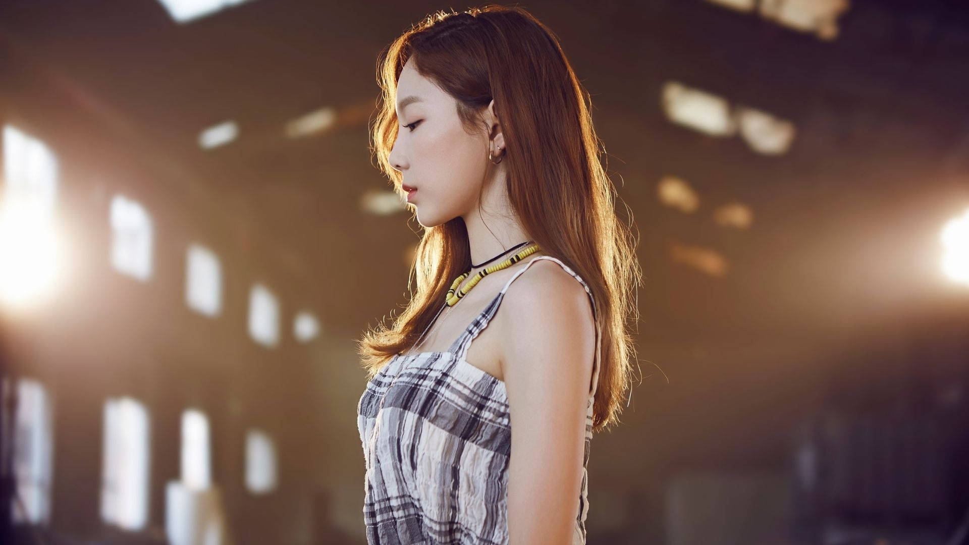 Taeyeon 桌布 壁紙 — 一位長棕色頭髮的年輕女性，在陽光普照的工業建築內側面站立，沐浴在高窗透進的溫暖金色光線中。