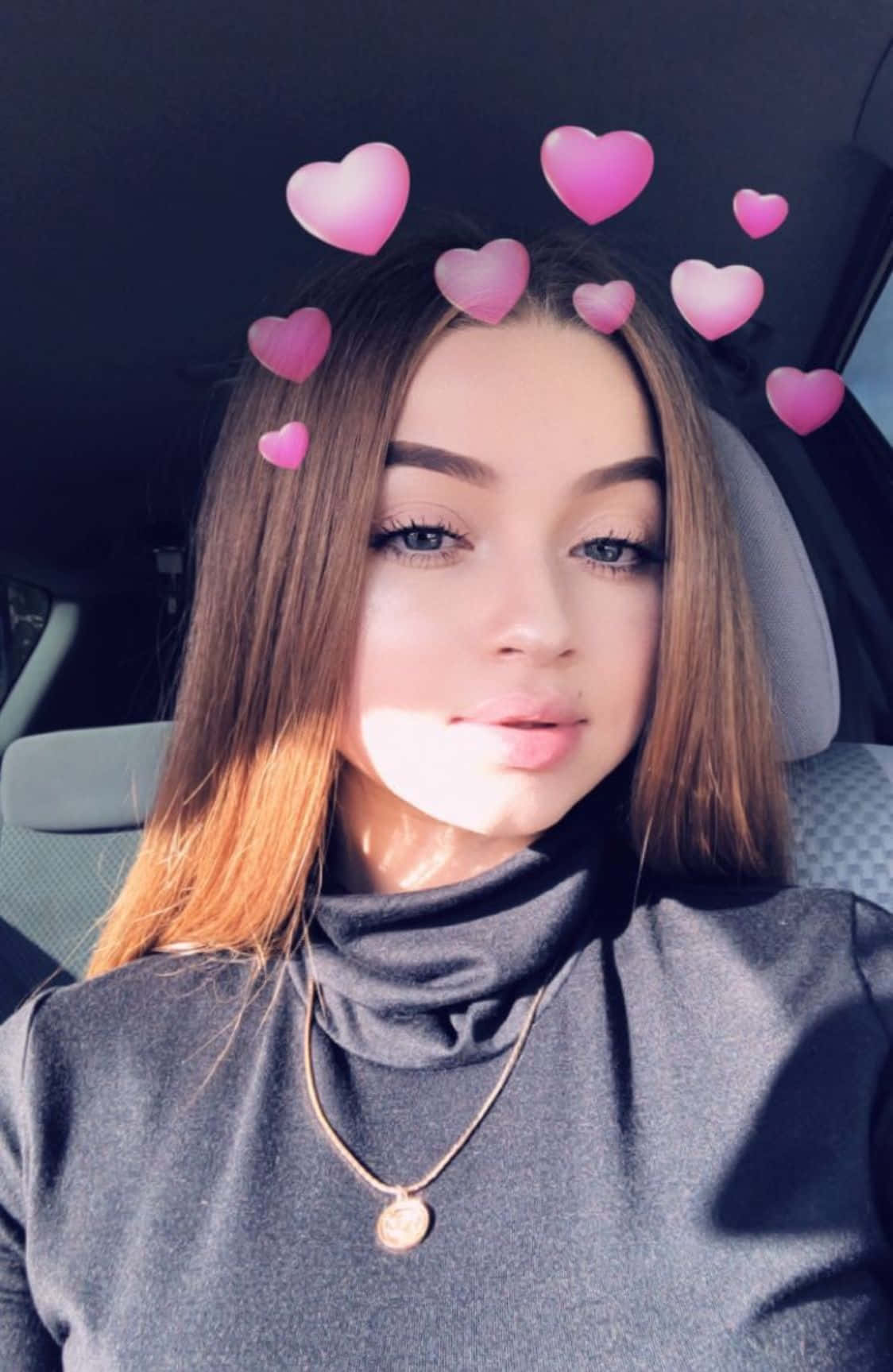 Snapchat 桌布 壁紙 — 一位長棕色頭髮的年輕女性坐在車裡，穿著灰色高領毛衣，粉紅色愛心表情符號漂浮在她的頭周圍。