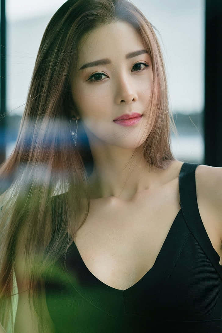 Cute Girl 桌布 壁紙 — 一位長棕髮、身穿黑色上衣的女性，透過模糊的綠色前景溫柔地凝視。