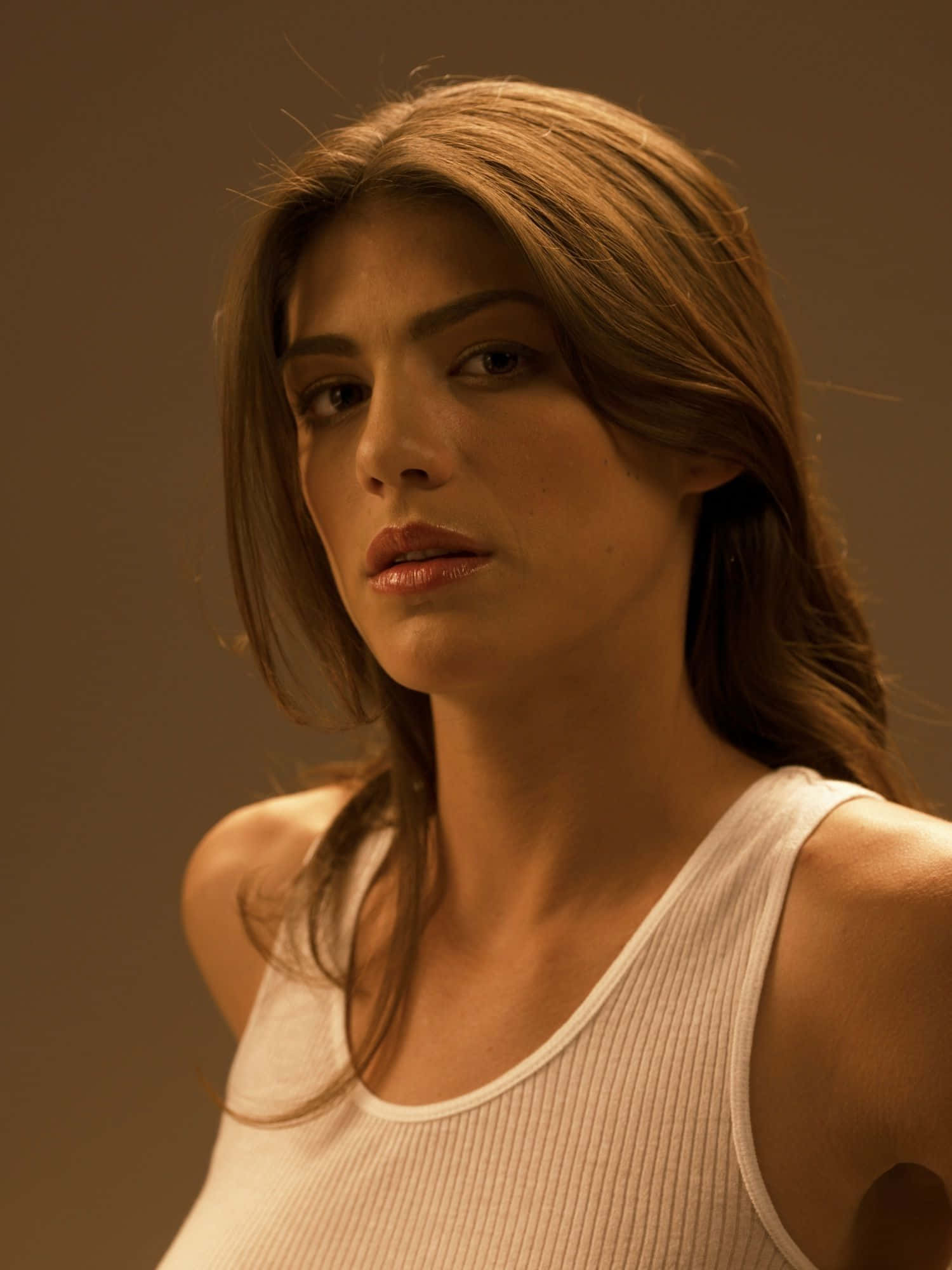 Genevieve Padalecki 桌布 壁紙 — 一位長棕髮女性在溫暖的攝影棚燈光下，穿著白色羅紋背心，背景為純棕色。