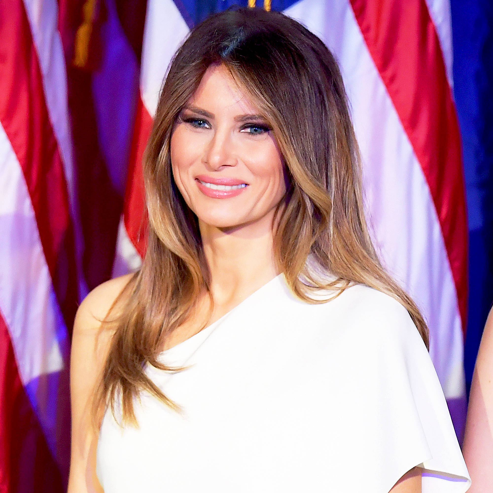 Melania 桌布 壁紙 — 一位長棕髮女性在美國國旗前穿著白色單肩裙溫暖微笑。